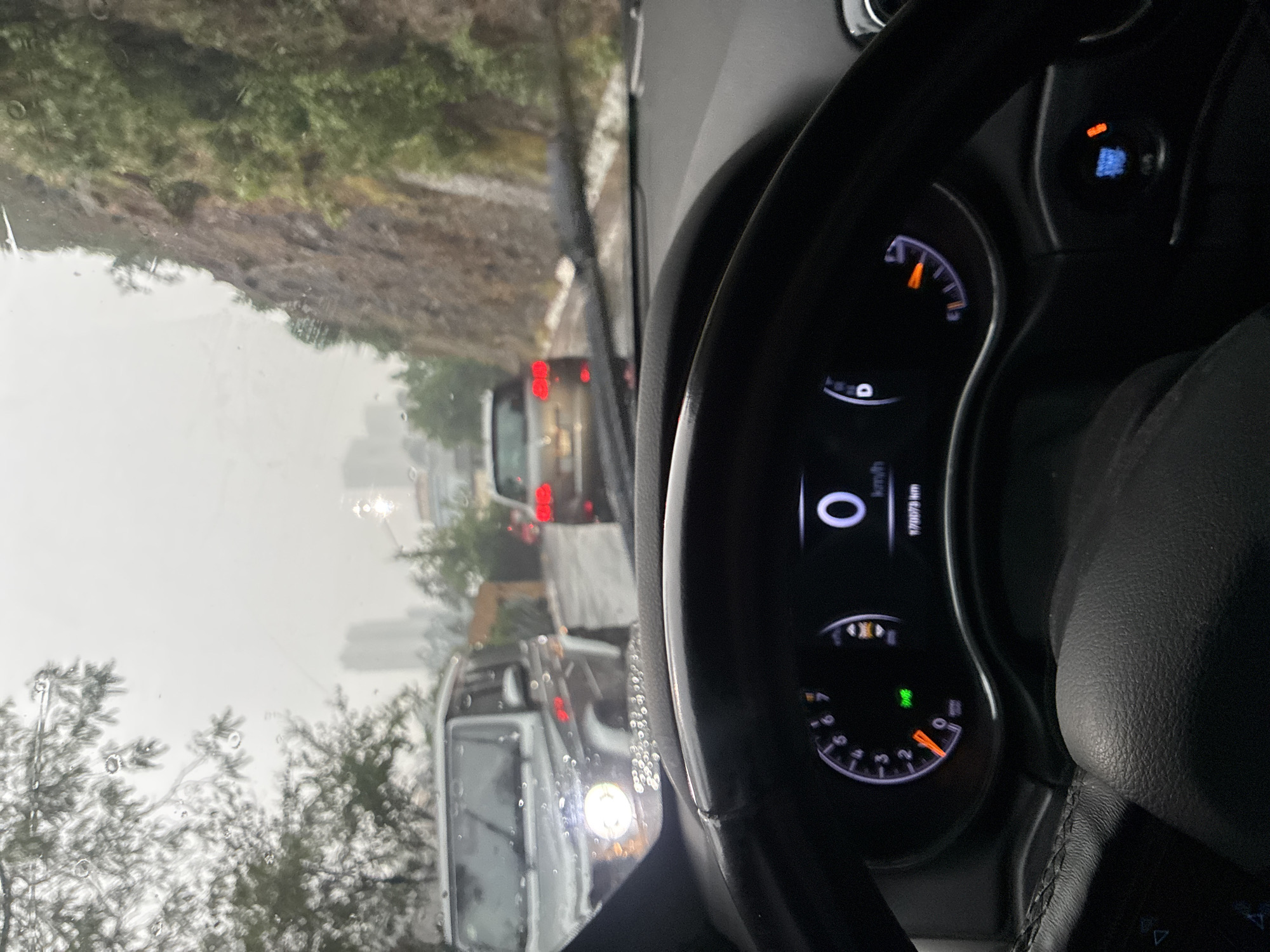 The image, taken from inside a stationary car, shows a view of traffic on a rainy day. The car's dashboard is visible, with the speedometer indicating 0 km/h. Through the windshield, other vehicles are seen in a line, including a white van with its headlights on and a red car further ahead. Trees and a hillside line the road on the left. In the distance, buildings, possibly part of the Mexico City skyline, are visible through the rain and mist. Based on the terrain and urban background, the location is likely on a road leading out of or into Mexico City.