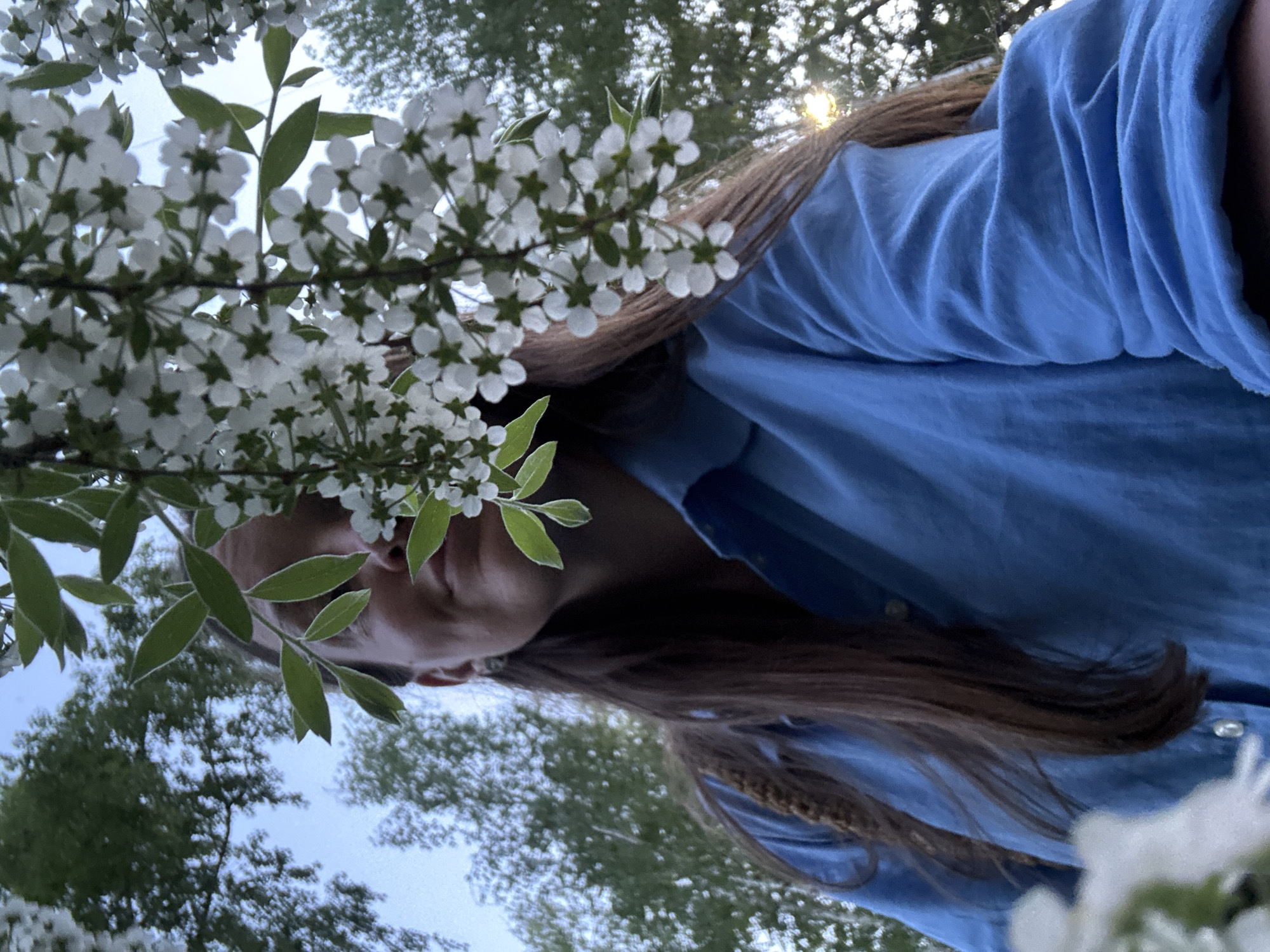 A low-angle, outdoor photograph captures a person in a blue shirt standing beneath a flowering tree branch. The individual's face is partially obscured by the leaves and blossoms, providing only a glimpse of their nose and mouth. Long, brown hair falls down along the person's shirt. The background reveals tree branches and the sky. The location cannot be confirmed as Chaikovsky, Russia, without further contextual information.