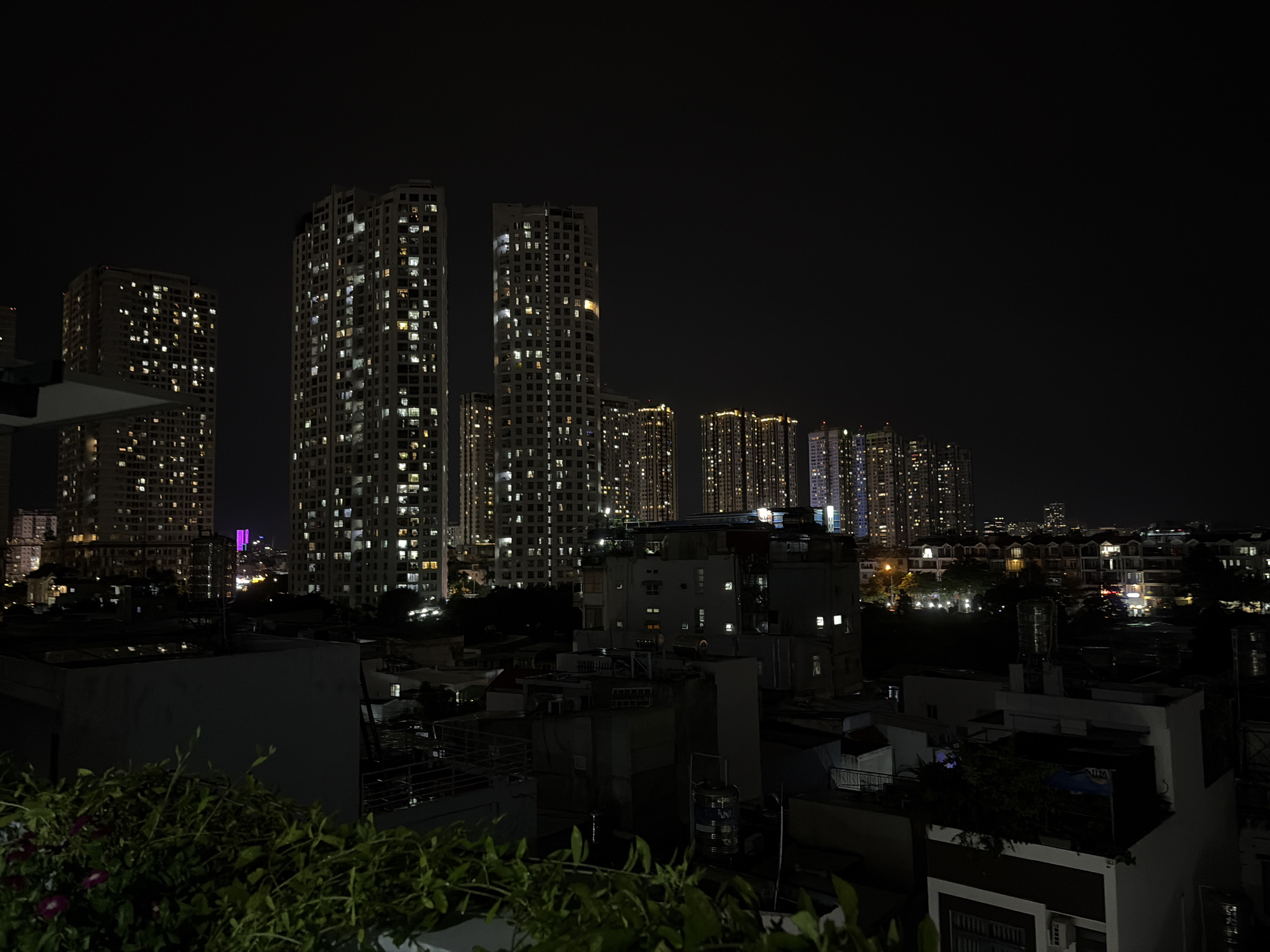 The image shows a nighttime cityscape in Thủ Đức, Vietnam, with a focus on illuminated high-rise buildings and lower-rise structures in the foreground. The sky is dark, indicating it is nighttime. Several tall apartment buildings are visible in the background, their windows lit with warm, yellow, and white lights, suggesting occupancy. Some of these buildings appear to have a grid-like pattern of illuminated windows. In the mid-ground and foreground, there are multiple lower buildings, some with visible windows and structures like balconies or rooftop terraces. In the very front, there is lush green foliage, likely from a plant or vine, obscuring a portion of the lower buildings. The overall scene depicts an urban environment at night, characterized by the contrast between dark skies and the glow of artificial lighting from residential and commercial structures.