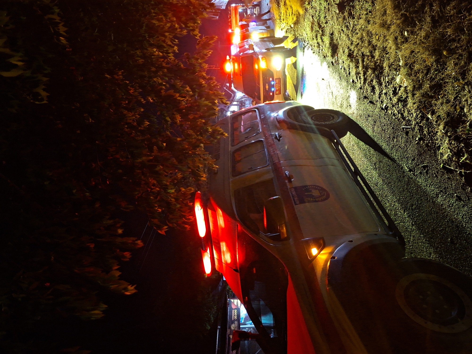 The image depicts a nighttime scene in Lhokdagang, Indonesia, with multiple emergency vehicles, likely ambulances or police cars, parked along a road. Red and blue lights from the vehicles illuminate the surroundings, casting a glow on the wet asphalt and the grassy verge. The foreground shows the side of a white emergency vehicle with red markings and lights on its roof. In the background, other similar vehicles are visible, as well as a person standing near one of them. Trees with dark leaves hang over the scene, their branches silhouetted against the illuminated sky. The overall impression is one of an ongoing incident or emergency response.
