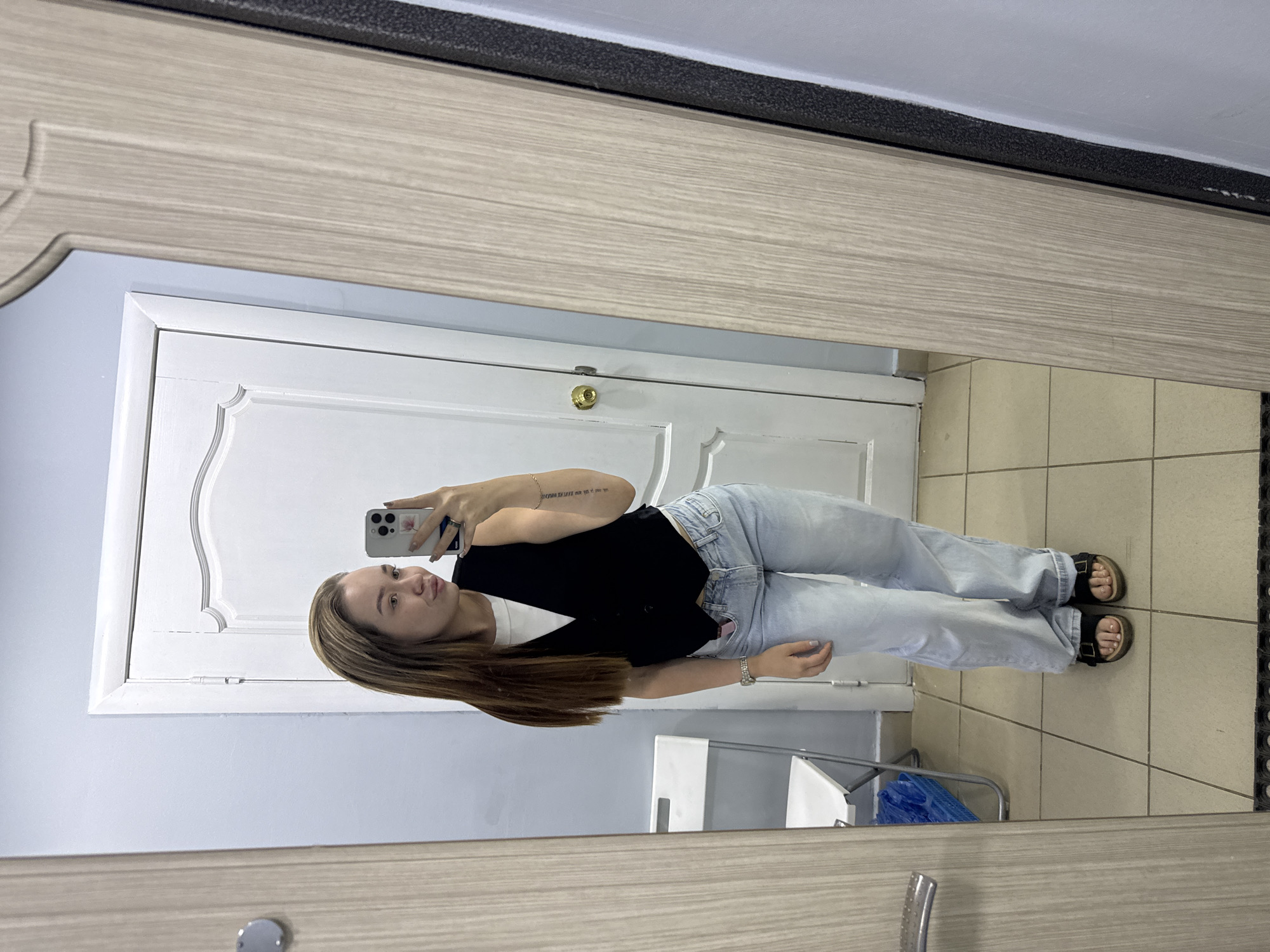 A woman is taking a selfie in a mirror. She is wearing a black top, light blue jeans, and sandals. She has brown hair. The mirror is framed in light-colored wood. Behind her is a white door and light blue wall. The floor is tiled in beige. In the background, there is a blue object. The location is inferred to be Chaikovsky, Russia.