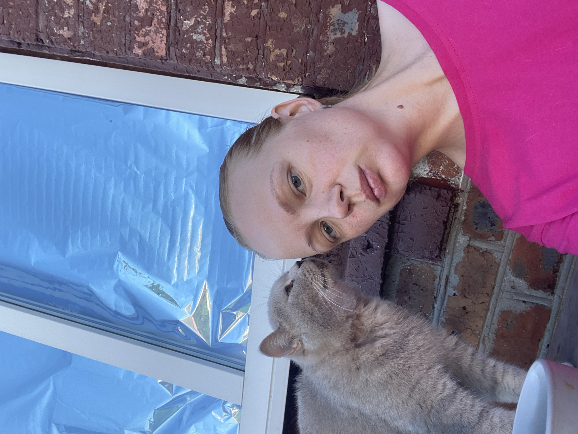 An outdoor, eye-level, medium shot captures a woman in a bright pink t-shirt and a light gray cat. The woman's head is tilted down and to her left, her gaze is directed slightly forward and to her left, and her lips are slightly parted. Her blonde hair is pulled back, and she has a small mole on her neck. The cat is positioned to her left, with its head turned towards her face, appearing to nuzzle her cheek. The cat is fluffy, with light gray fur.
The background consists of a brick wall with peeling paint, a white window frame, and a blue, reflective surface that appears to be a window covering or plastic sheeting. The setting suggests an outdoor, possibly urban, environment, consistent with a location in Kisliakovskaya, Russia. The lighting indicates it is daytime. The composition is a selfie, with the woman and cat as the primary subjects.