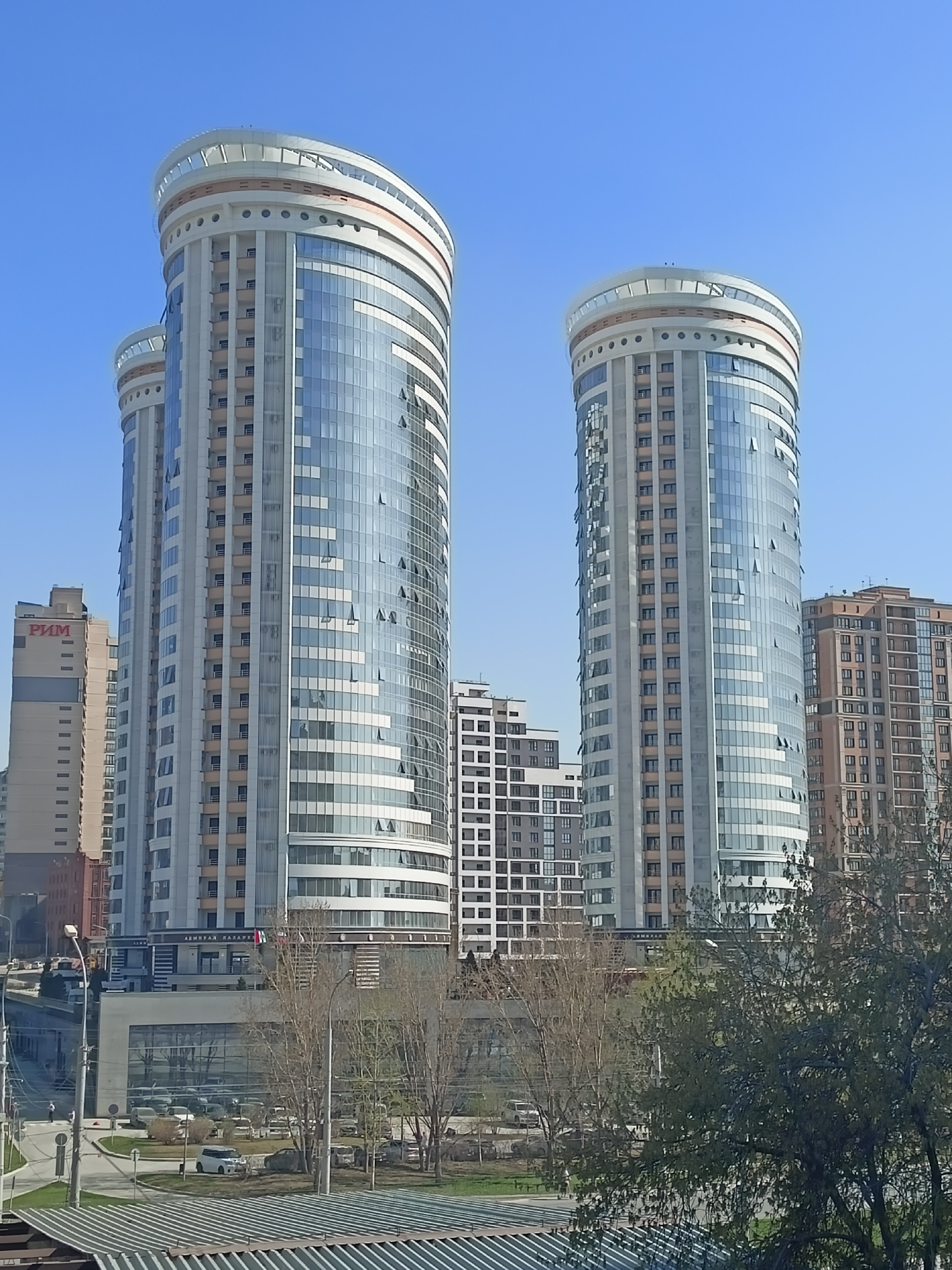 The image shows two cylindrical skyscrapers in Novosibirsk, Russia, under a clear blue sky. The buildings have a distinctive design with glass facades and decorative elements at the top. A smaller building is visible between the two larger structures. Trees are in the foreground, partially obscuring the view of the street and buildings below. A building with the word "РИМ" (RIM) on the side is visible to the left of the left tower. Cars are parked in the area below.