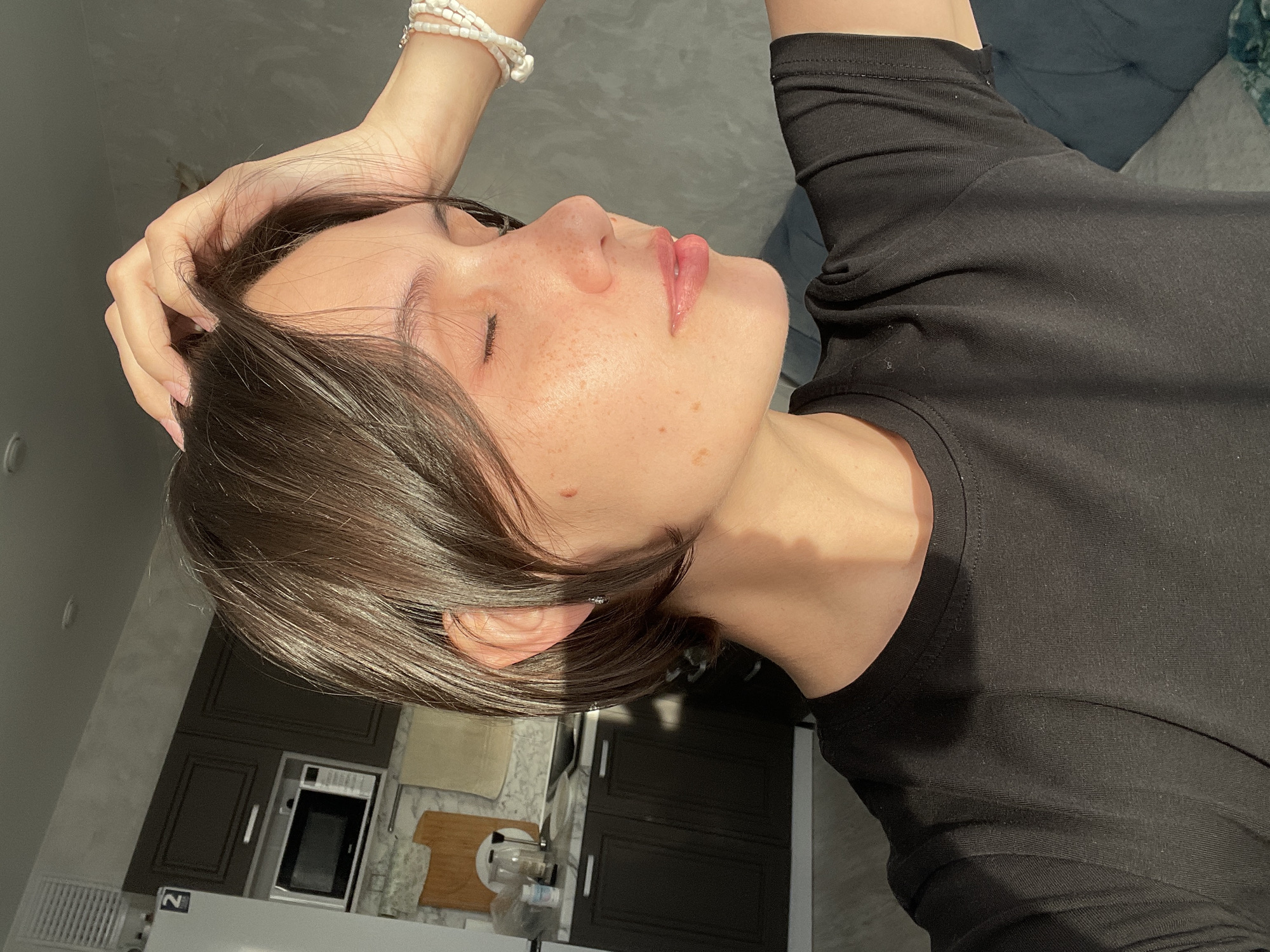 A brunette woman lies on her back, seemingly indoors, with her right arm bent above her head. She wears a black, long-sleeved shirt. Her eyes are closed and her skin appears to have freckles. She is wearing a white beaded bracelet on her right wrist. The location is a modern kitchen, including dark cabinetry, white countertops, a microwave, a stove and kitchen accessories. It is impossible to determine whether the location is in Izhevsk, Russia from the image.