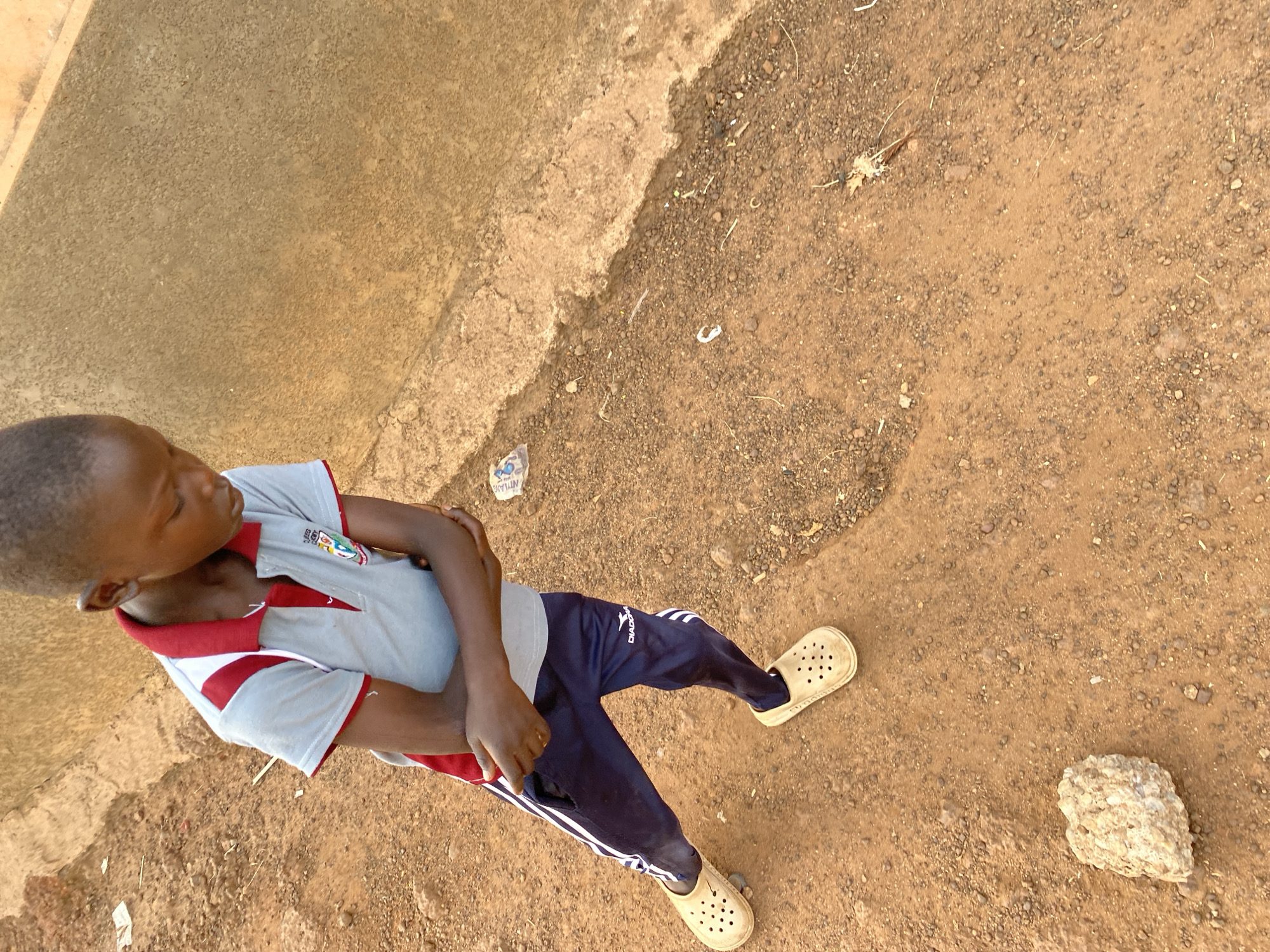 An overhead shot captures a young boy standing outdoors in Bukuru, Nigeria. He is positioned slightly to the left of the frame, looking down and to his left with his eyes cast downwards. His arms are crossed in front of him.
The boy has short, dark hair. He is wearing a light grey short-sleeved polo shirt with a red collar and red and white striped trim on the sleeves. A small, colorful crest-like emblem is visible on the left chest of his shirt. He wears dark blue track pants with two prominent white stripes running down the outer sides of the legs. A white "DIADORA" logo is clearly visible on his left thigh. On his feet, he wears light beige, perforated clogs.
The ground beneath him consists of two distinct surfaces: a smoother, light brown concrete or paved area occupies the left side of the frame, while the larger right portion is a rougher reddish-brown dirt and gravel surface. This dirt ground is strewn with small stones, a few dry leaves, and scattered pieces of litter, including a small crumpled plastic bag near the bottom left edge. A distinct light-colored, irregularly shaped rock lies on the dirt ground to the right of the boy's feet. The natural lighting suggests it is daytime. The scene depicts a casual, candid moment in an outdoor setting.