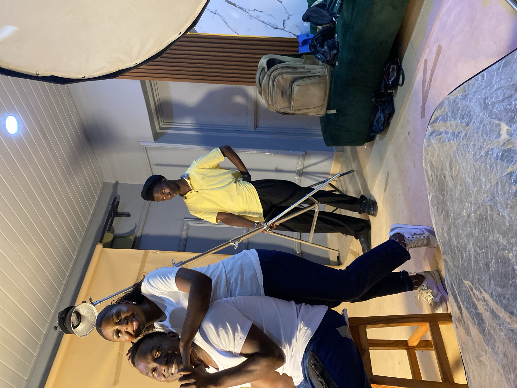 The image depicts an indoor scene, likely a studio setting, in what appears to be Bukuru, Nigeria based on the context provided. Three people are visible: two are posing closely, with the woman in a white shirt leaning over a man, also in a white shirt, who is smiling at the camera. A third person, a man wearing a yellow shirt and black pants, stands in the background, his hand on his hip, seemingly overseeing the setup.
The location is equipped with photography equipment. Light modifiers, including a large softbox above the posing subjects and another diffused lighting element to the side, indicate the scene is prepared for a photo shoot. A photography backdrop is set behind the posing subjects. Other objects present include a light stand being managed by the man in yellow, a wooden chair, and bags piled on a sofa in the background. The interior walls have different styles of paneling and paint.