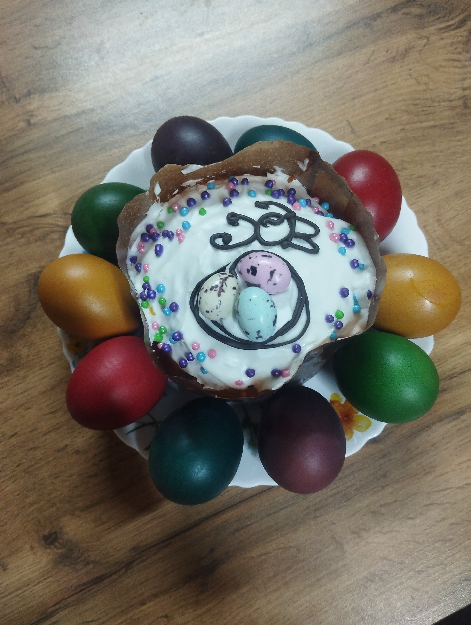 The image shows a top-down view of an Easter cake and dyed Easter eggs on a plate. The plate rests on a wooden surface. The Easter cake has white frosting, multicolored sprinkles, and a chocolate decoration that includes a drawing of eggs with "XB" inscribed above them. The plate is bordered by twelve dyed eggs in varying colors, including red, green, yellow-orange, dark green, and purple. The plate itself is white with a scalloped edge and a floral pattern. The wooden surface appears to be a tabletop, with a wood grain pattern visible. The location is Grishanki, Russia. There are no people visible in the image.