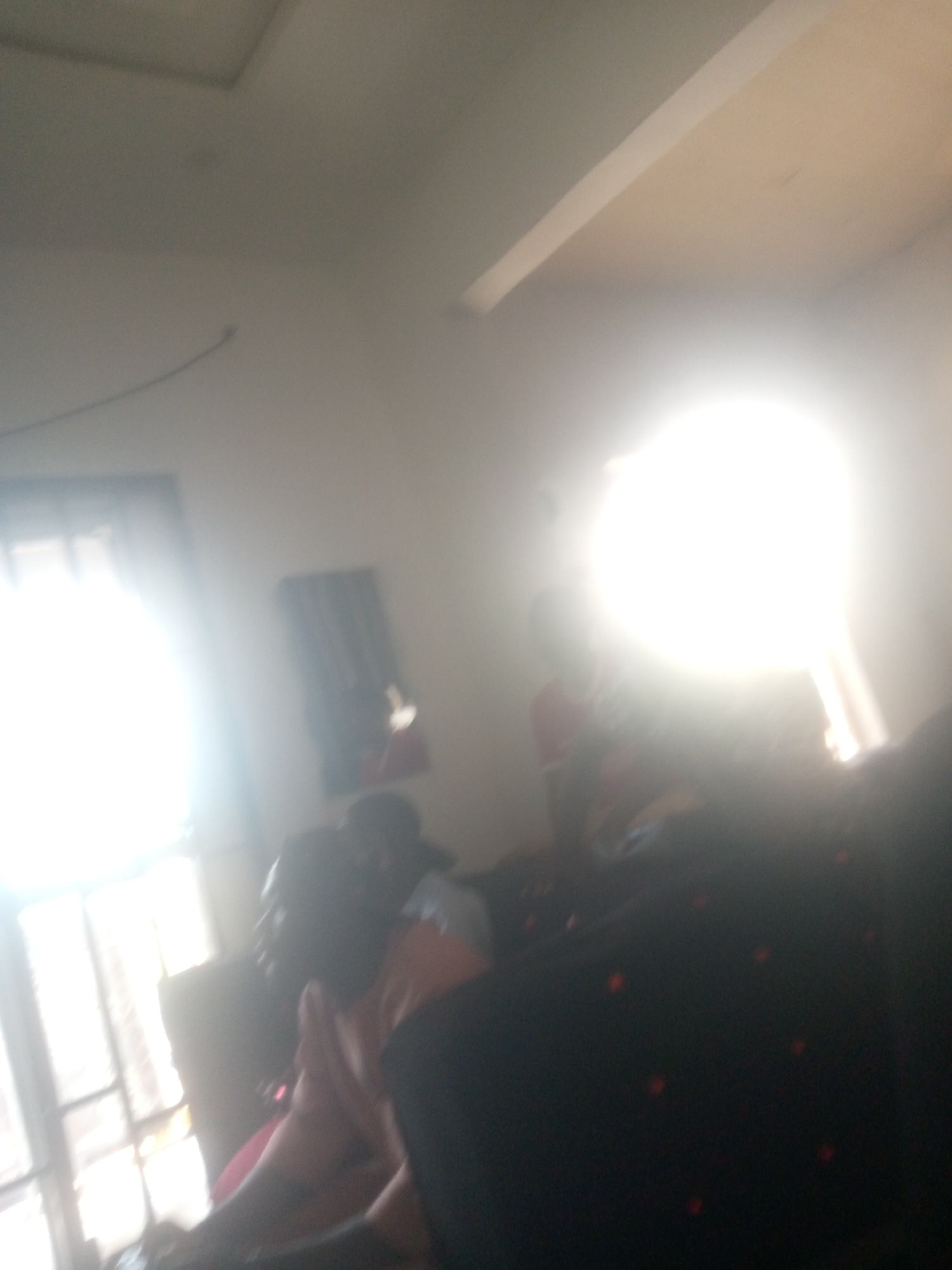 The image appears to be an indoor scene, possibly a living room, in Jalingo, Nigeria. The lighting is extremely bright, suggesting a strong light source is present, potentially a flash or direct sunlight, which overexposes much of the image. In the foreground, there's a dark upholstered surface, possibly a sofa, with several red circular patterns visible. Behind this, a blurred figure of a person is partially visible, seated and facing away from the camera. To the left, a brightly lit window with a grid pattern is visible, suggesting it might be a casement window. A curtain or blinds are partially drawn. Further back, another blurred figure, possibly of a woman, is seated. The walls are plain, and a portion of the ceiling is visible, appearing to be a dropped ceiling with a linear light fixture. The overall impression is that of a casual indoor setting, but the extreme overexposure makes specific details difficult to discern.