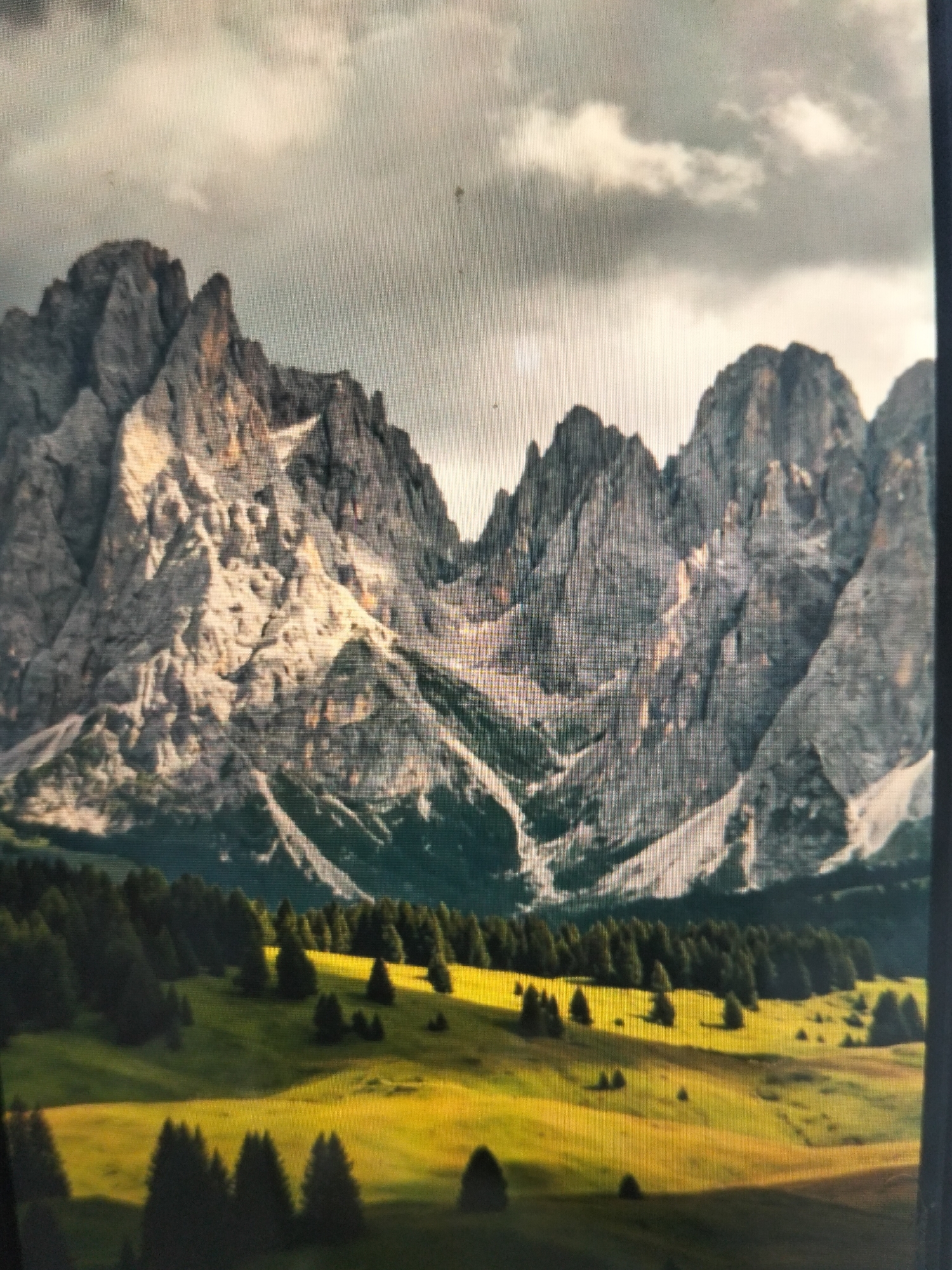 The image depicts a dramatic mountain landscape under a cloudy sky. Jagged, rocky peaks dominate the background, with sunlight illuminating sections of their steep faces, creating strong contrasts between light and shadow. Patches of snow are visible on the higher slopes. In the mid-ground, a dense band of dark green pine trees forms a line across the base of the mountains. In the foreground, rolling hills covered in bright, sunlit green and yellow grass are dotted with scattered evergreen trees. The overall scene is one of natural grandeur and isolation.
This image does not depict the city of Payagaboh, Indonesia.