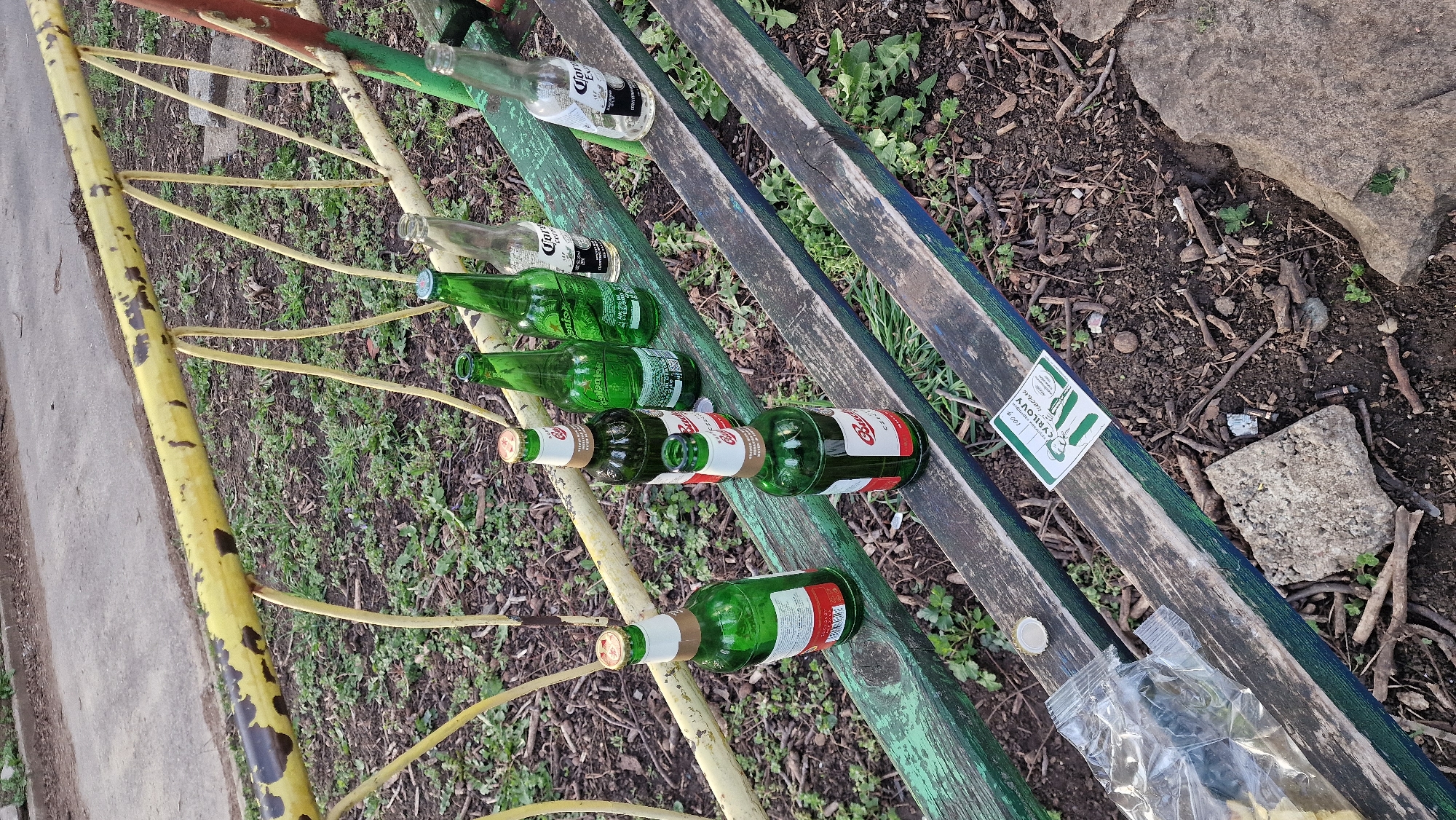 The image shows a park bench in what appears to be a neglected state. Several empty beer bottles, including Corona and other green glass bottles, are scattered along the length of the bench. There is a sticker with the word "CYRILOVY" visible on one of the wooden planks. Debris such as a plastic bag, a bottle cap, twigs, leaves, and a small rock are also present on and around the bench. The bench itself is painted with green and yellow paint that is peeling. The background consists of dirt, grass, and more vegetation.