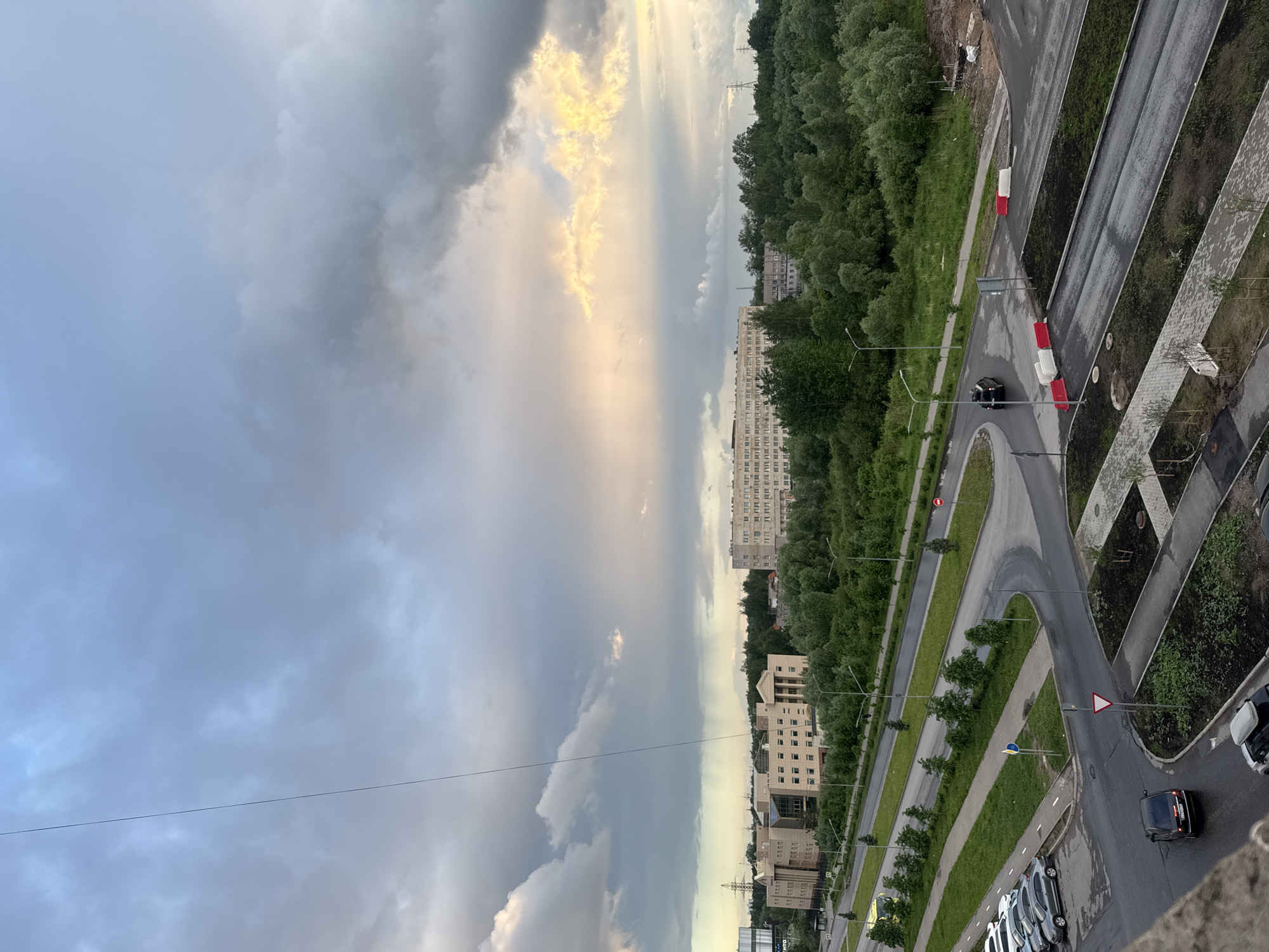 The image shows an aerial view of a section of Veliky Novgorod, Russia. The sky is dominated by storm clouds with a patch of sunlight breaking through. Below the clouds, a mix of trees and buildings are visible. The architectural style of the buildings appears typical of Soviet-era construction. A road network is visible, including cars parked on the side of the street and one driving on the street. The road layout includes a small roundabout or turning circle. There are also landscaped areas with grass and small trees alongside the road.