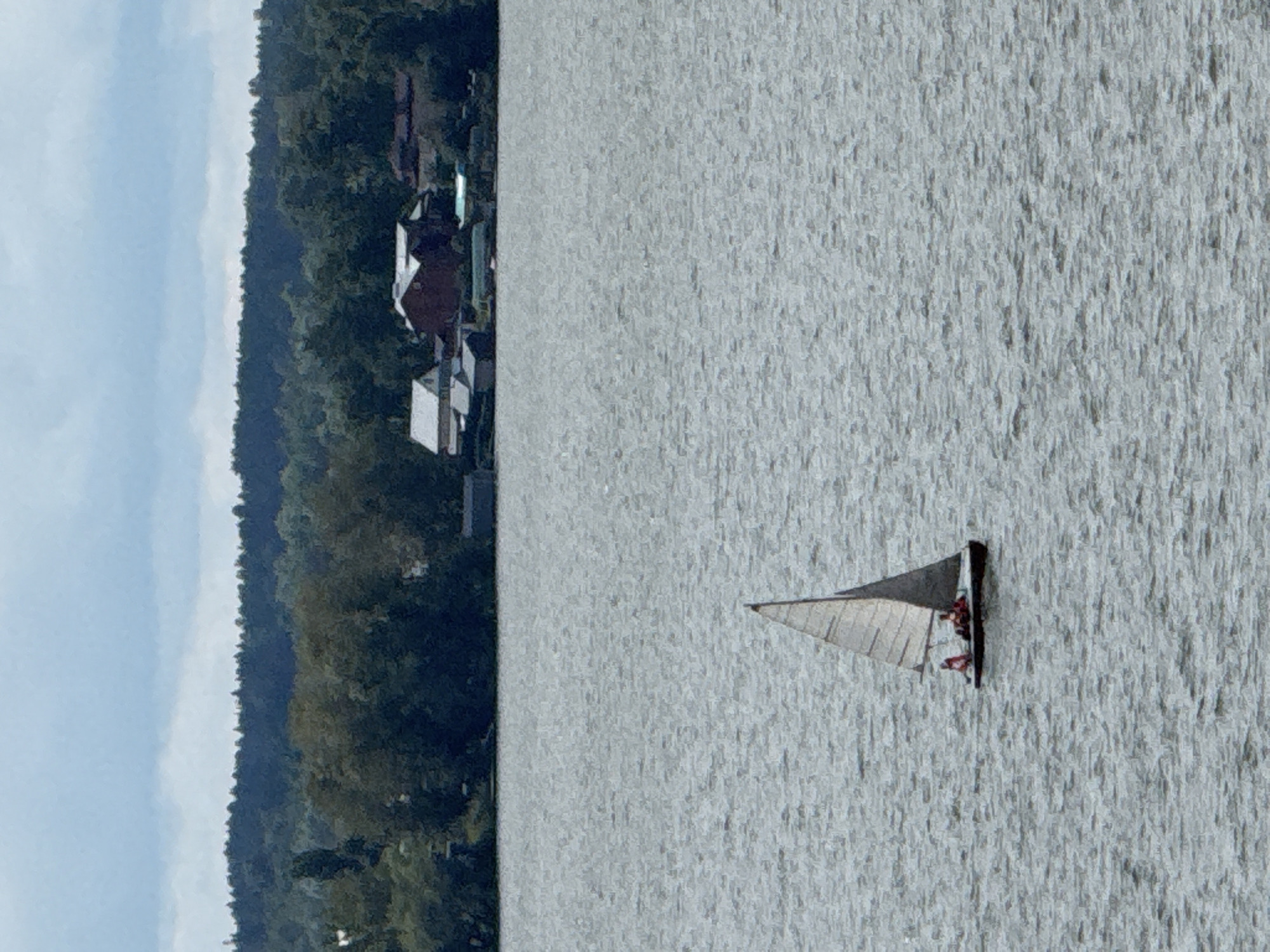The image shows a wide body of water, likely the Kama River, on an overcast day. In the foreground, a small sailboat with two individuals on board is sailing. The boat has a white sail. In the background, a densely wooded shoreline is visible, with trees forming a dark green line against the sky. A few buildings, appearing to be houses and possibly other structures, are situated along the shoreline amidst the trees. The overall impression is of a peaceful, natural setting with signs of human habitation near the water's edge. The image is undated and the specific location within Votkinsk, Russia, is not discernible from the visual information alone, though the Kama River flows through the region.