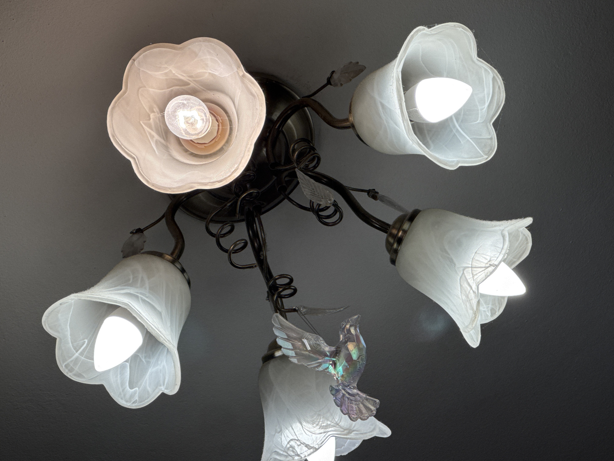 A ceiling light fixture hangs from a dark ceiling. The fixture has a dark metal frame with decorative elements that resemble leaves and vines. There are five lamp shades, four of which are illuminated. Four shades are bell-shaped and made of white, frosted glass with swirling patterns. One of these shades is also decorated with a small, iridescent bird figurine. The fifth shade, located at the top left, is more open and flower-shaped, also made of frosted glass, and contains an exposed light bulb. The overall lighting is dim, with the light from the fixture being the primary source. This image was taken indoors, likely in a residential setting. There is no information to suggest this location is in Tallinn, Estonia.
