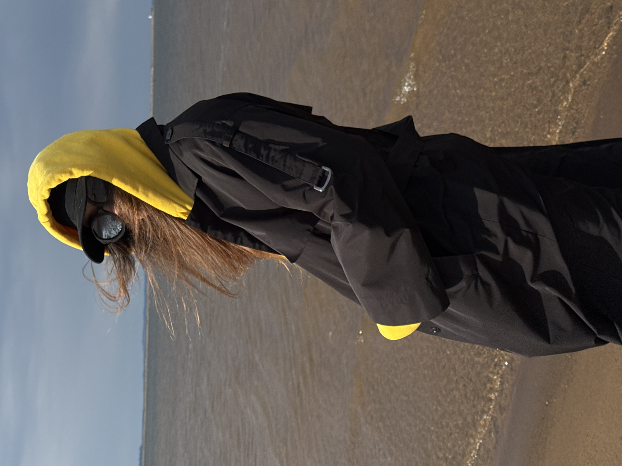 An individual with long, brown hair is depicted standing at the edge of a body of water under a clear sky. The person is dressed in a black jacket featuring a prominent yellow hood. A black baseball cap is worn beneath the hood, and dark, round sunglasses obscure their face. The person appears to be looking downwards or leaning slightly towards the water.
The setting is a shoreline, likely a riverbank given the location context of Chaikovsky, Russia (Kama River). The water is calm, with gentle ripples visible near the edge, and the shore consists of sand or fine pebbles. The sky above is a light blue, indicating fair weather during the daytime, possibly morning or late afternoon given the soft lighting. No specific activity beyond standing by the water is clearly discernible. There is no visible text in the image.