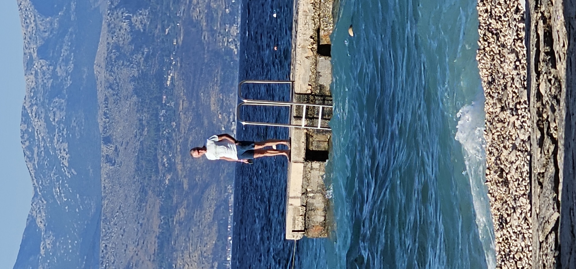 An older man with short gray hair, wearing a white t-shirt with dark text and dark blue shorts, stands on a concrete pier. He is holding a dark object in his right hand and has his left hand in his pocket. The pier has a metal ladder leading into the turquoise water. The water is choppy, with waves breaking on a pebble beach in the foreground. In the background, there are blue mountains with some vegetation and scattered buildings. The sky is clear and blue. The scene appears to be in Supetar, Croatia.