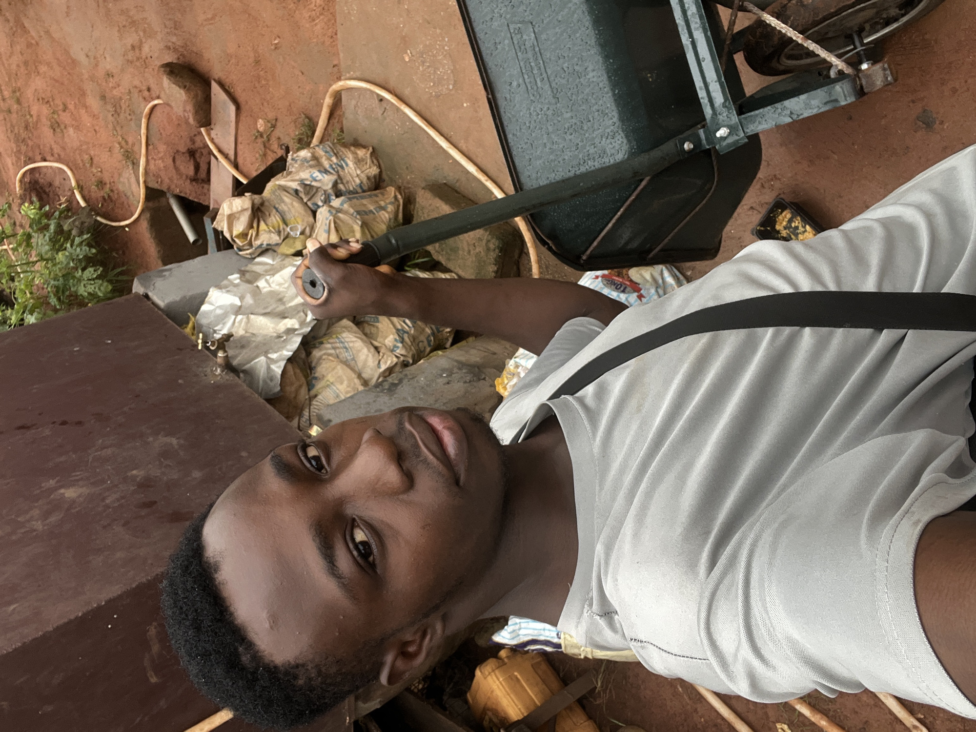 A young Black man is taking a selfie outdoors in what appears to be a somewhat cluttered or work-in-progress environment. He is wearing a light gray t-shirt with a black strap across it. His hair is short and black. He is holding the handle of a dark green wheelbarrow with his right hand. The wheelbarrow is positioned to his right and has a single wheel.
The background is composed of various elements. To the left and behind him is a large, dark brown, possibly metal or concrete structure. In the mid-ground, there are stacks of large, light-colored bags, some with visible text that is difficult to read. A yellow plastic container is also visible among these bags. A light-colored hose is present, snaking across the reddish-brown ground and partially obscured by rocks and a concrete structure. Some green foliage is visible in the upper right of the image. The ground is reddish-brown, suggesting it is made of clay or laterite soil, common in many parts of Nigeria.
The overall impression is that the man might be in a construction site, a farm, or a yard where materials are being stored or moved. The presence of the wheelbarrow and bags points towards manual labor. The image suggests it could be Nkpor, Nigeria, given the characteristic red earth and the general visual context.