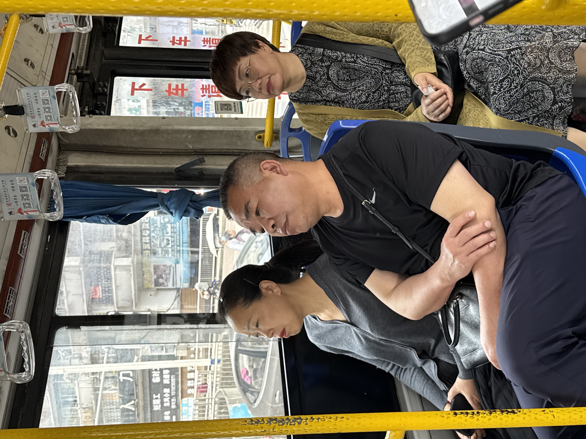 The image captures a scene inside a public bus. There are three passengers visible: an older woman wearing glasses and a patterned dress, a man in a black T-shirt, and a woman in a grey shirt. They are seated on blue seats with yellow safety bars in the bus. Through the bus window, buildings with Chinese characters are visible. The Chinese characters suggest the location is in China. Based on the characters visible, particularly the "Jiangjin" (江津) character, the location is most likely in the city of Jiangjin.