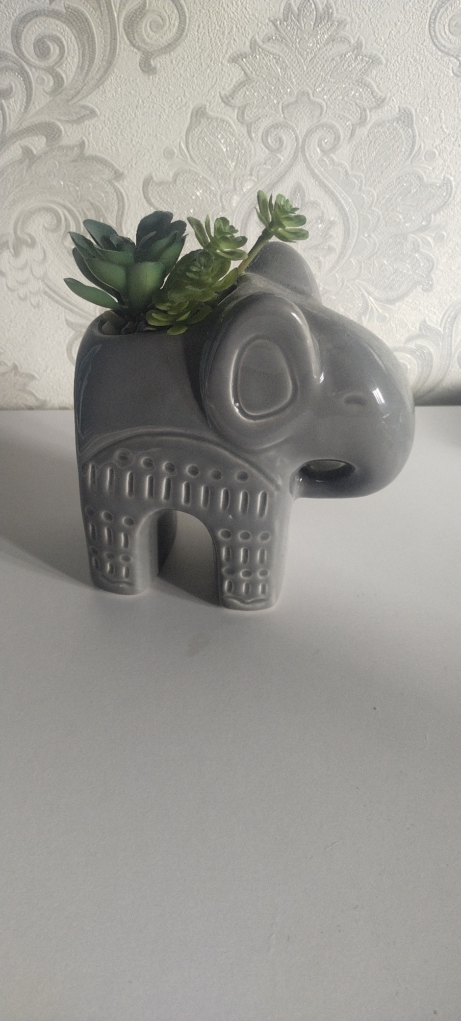 A grey ceramic elephant planter sits on a white surface in front of a textured white wallpaper with a damask pattern. The planter has carved details on its body, resembling eyes and decorative patterns on its legs and sides. It contains small green succulent plants and tiny pebbles. The lighting casts a soft shadow from the planter onto the white surface.