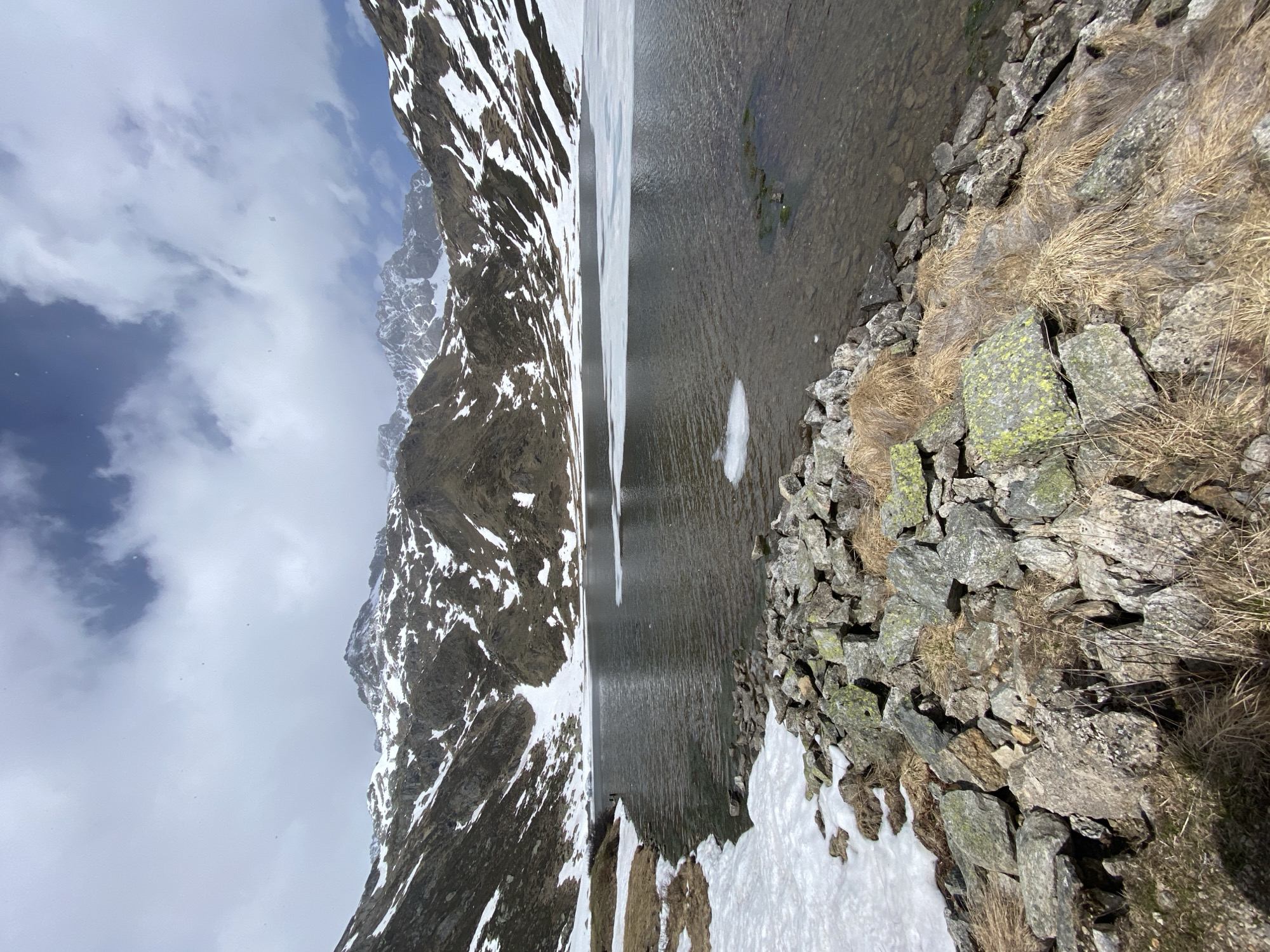 The image shows a landscape near Baceno, Italy. A lake occupies the central part of the scene, reflecting the surrounding mountains and sky. The mountains are partially covered in snow, with rocky terrain visible underneath. The shoreline consists of rocks and patches of snow and dead grass. The sky is overcast with clouds.