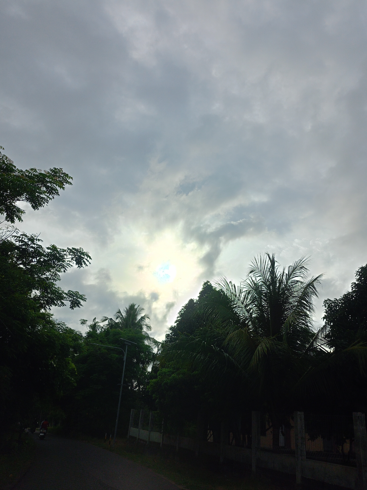 The image shows a cloudy sky above a road in Meunasahteungkudigadong, Indonesia. Trees are visible along the roadside, with a few palm trees interspersed. A fence runs alongside the road. The sun is obscured by the clouds, but its position is indicated by a bright patch in the sky. A person on a motorcycle can be seen on the left side of the road.