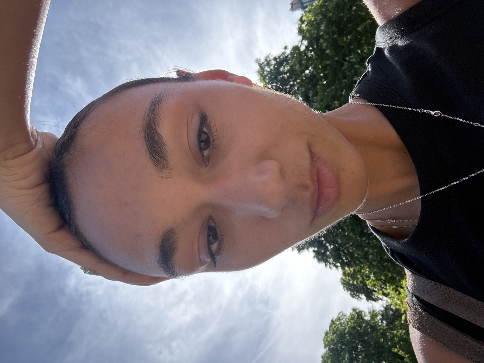 A close-up, upward-facing selfie shows a person with light skin, dark eyebrows, and brown eyes. The person has a hand raised, partially obscuring their forehead. They are wearing a black t-shirt and a delicate silver necklace. In the background, the sky is visible, with white clouds. Trees are also visible in the background. The location appears to be an outdoor area in Paris, France.