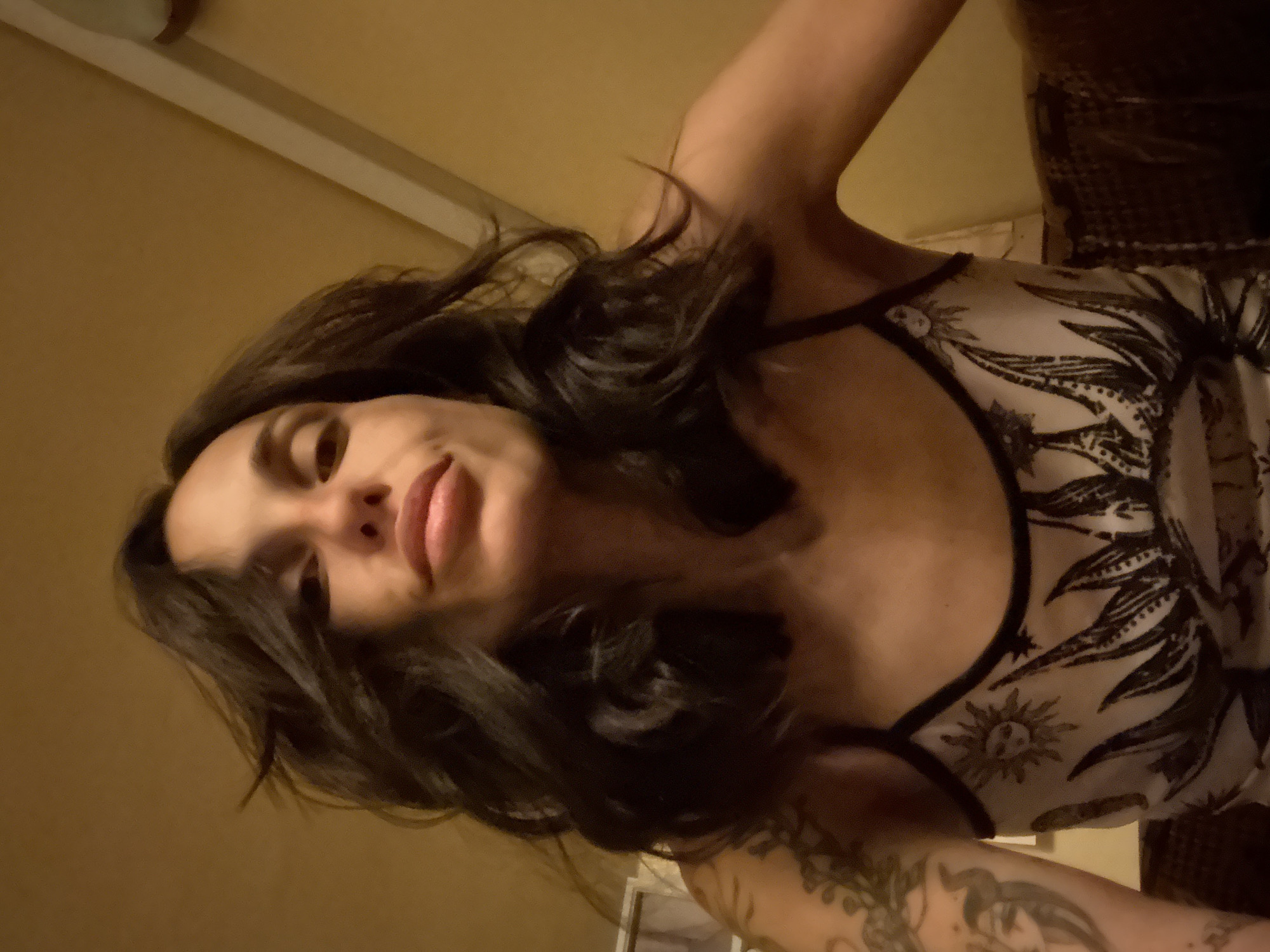 The image is a close-up, low-angle selfie of a woman with dark, wavy hair. She is looking directly at the camera with a neutral expression. Her lips are slightly parted. She is wearing a light-colored tank top with a black trim, featuring a graphic print of a sun and agave plants. Tattoos are visible on her left arm. The background appears to be an indoor setting with warm-toned walls and ceiling beams, suggestive of a rustic or bohemian interior. There is no discernible information within the image to indicate a specific location in Pleven, Bulgaria.