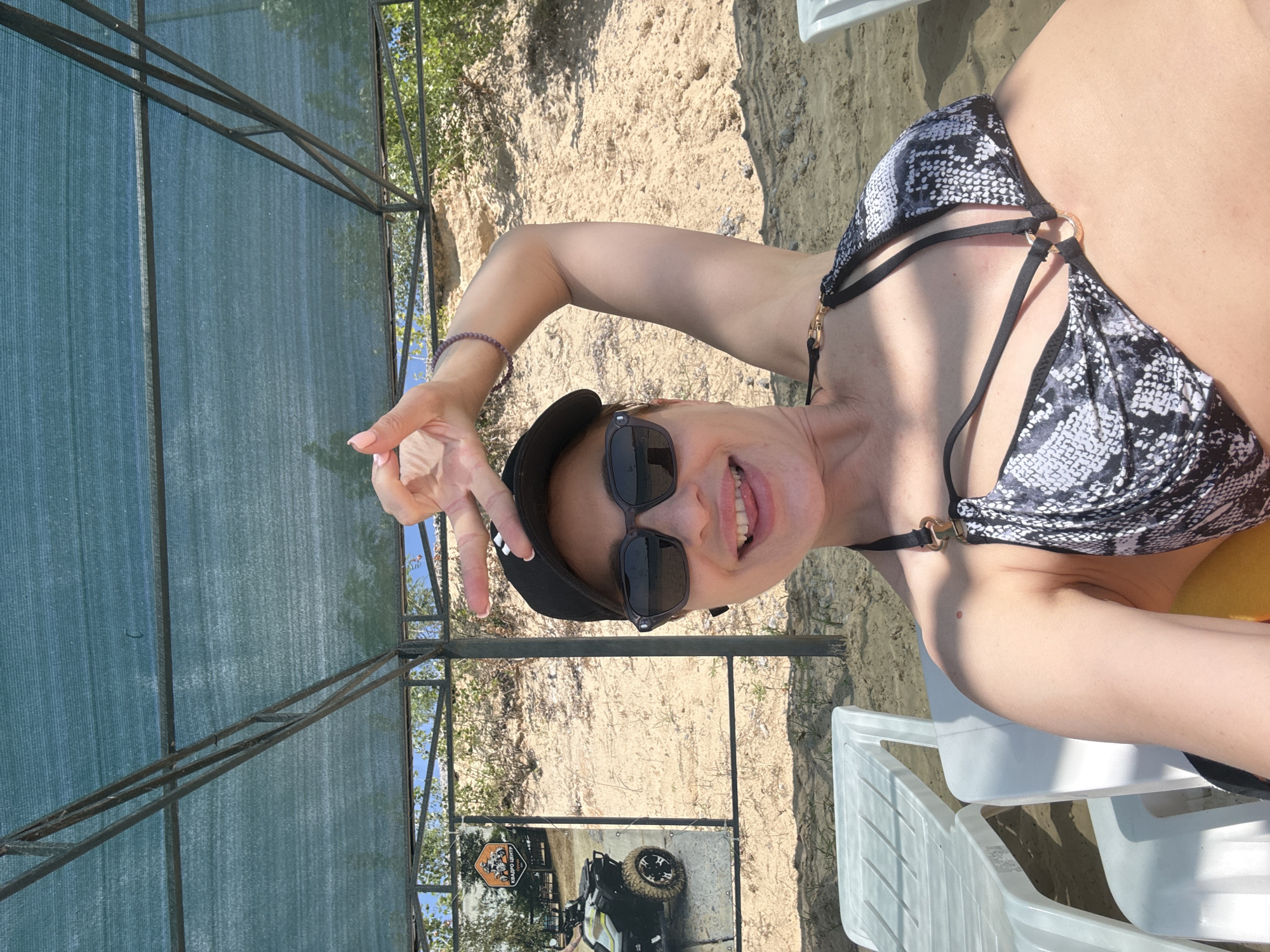 The image features a woman wearing a black and white snakeskin-patterned bikini top, a black baseball cap, and sunglasses. She is smiling and making a peace sign with her fingers. She is seated on a white plastic chair in an outdoor setting, likely a beach or resort area, indicated by the sand and vegetation in the background. To her left, a green mesh covering supported by a metal frame is visible. A poster advertising an ATV center is also present, featuring a picture of an ATV and text that reads "АГРО ЦЕНТР". The lighting suggests it is daytime, with bright sunlight casting shadows. The overall mood is cheerful and relaxed.
