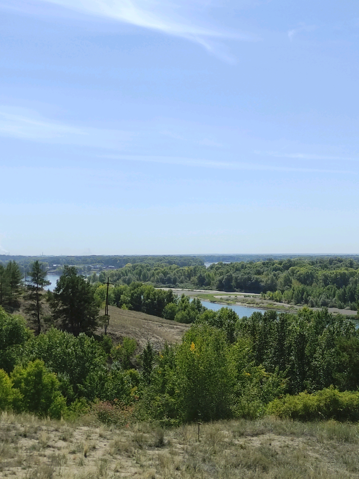 The image captures a panoramic view of a river flowing through a densely forested area under a clear blue sky. The foreground consists of a sandy, dry hillside covered with sparse, dry grass and some small, low-lying green bushes. Moving further into the scene, the terrain slopes down towards the river, with a section of drier, brownish earth interspersed with patches of green vegetation. Tall, mature trees with lush green foliage dominate the middle ground, forming a thick canopy that lines the riverbanks. A calm, wide river with a light blue hue meanders through the landscape, reflecting the sky. In the distance, the landscape transitions to a hazy horizon of trees, suggesting a vast expanse of forest. A lone power pole with wires is visible on the hillside in the middle ground, and a faint industrial silhouette can be seen on the far horizon. The overall scene is one of natural beauty and tranquility, characteristic of a rural or semi-rural environment. The location is identified as Glukhovo Rural District, Kazakhstan.