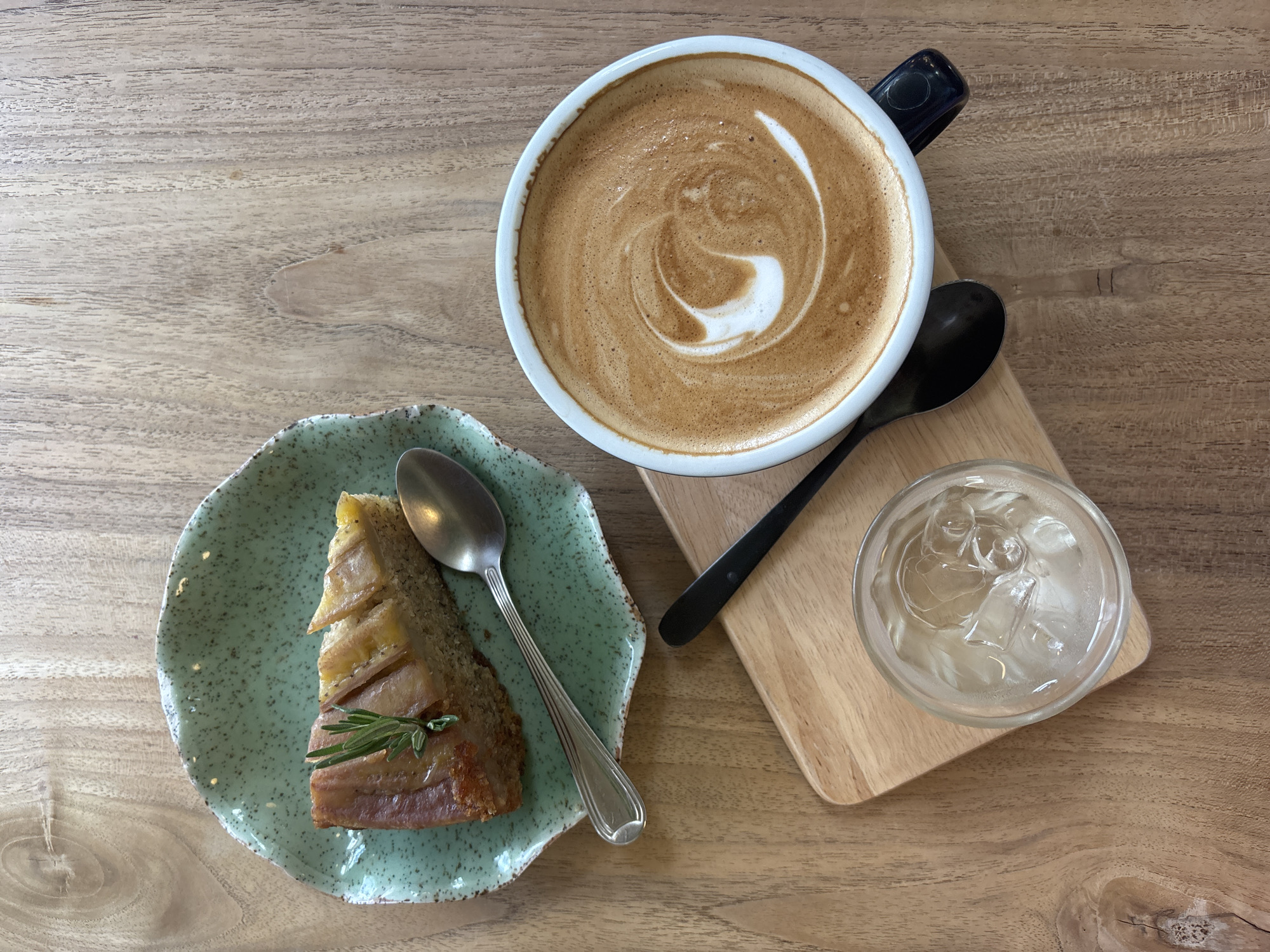 A top-down view on a wooden table presents a culinary still life, seemingly indoors in Choeng Thale, Thailand. A slice of cake, garnished with a sprig of rosemary, rests on a speckled green plate with a spoon alongside it. Adjacent to the plate, there is a large, white mug filled with latte or cappuccino adorned with latte art, accompanied by a black spoon. A wooden board sits close by, holding a glass filled with ice and water. The table's surface appears to be a light-colored wood with a visible grain.