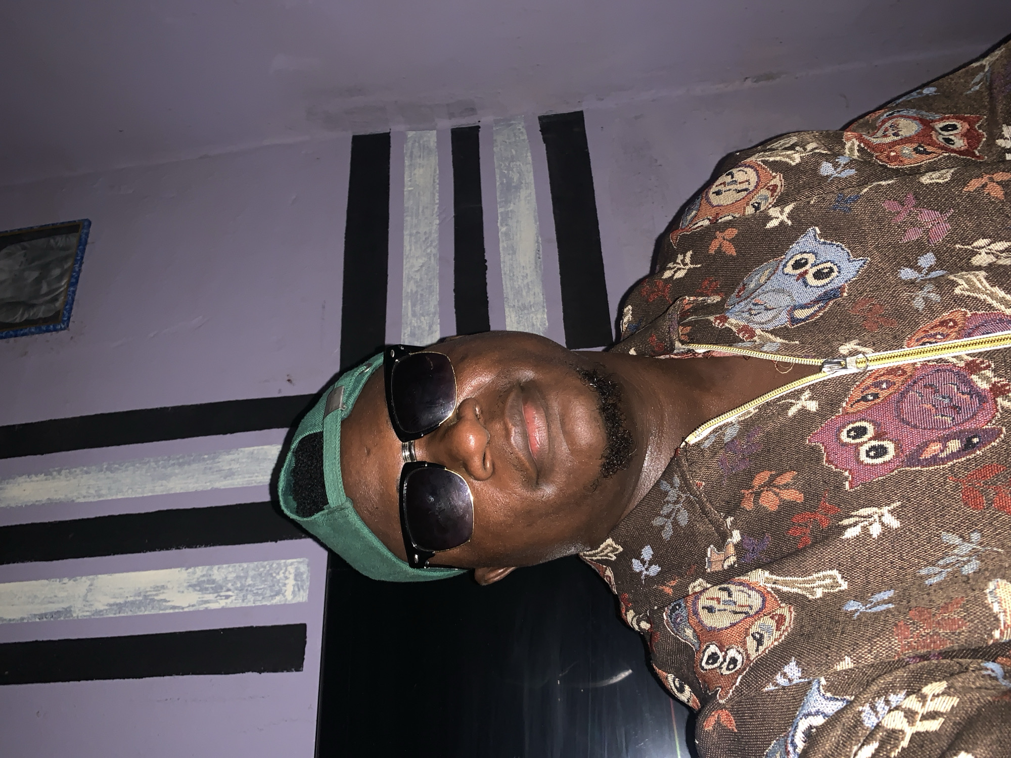 A man is seen from the chest up, wearing a green baseball cap and dark sunglasses. He is also wearing a brown, owl-patterned jacket with a yellow zipper. The background features a light purple wall with alternating black and white vertical stripes. A framed picture hangs on the wall to the left. It is impossible to determine the location in the city of Akwanga, Nigeria, from this image alone.