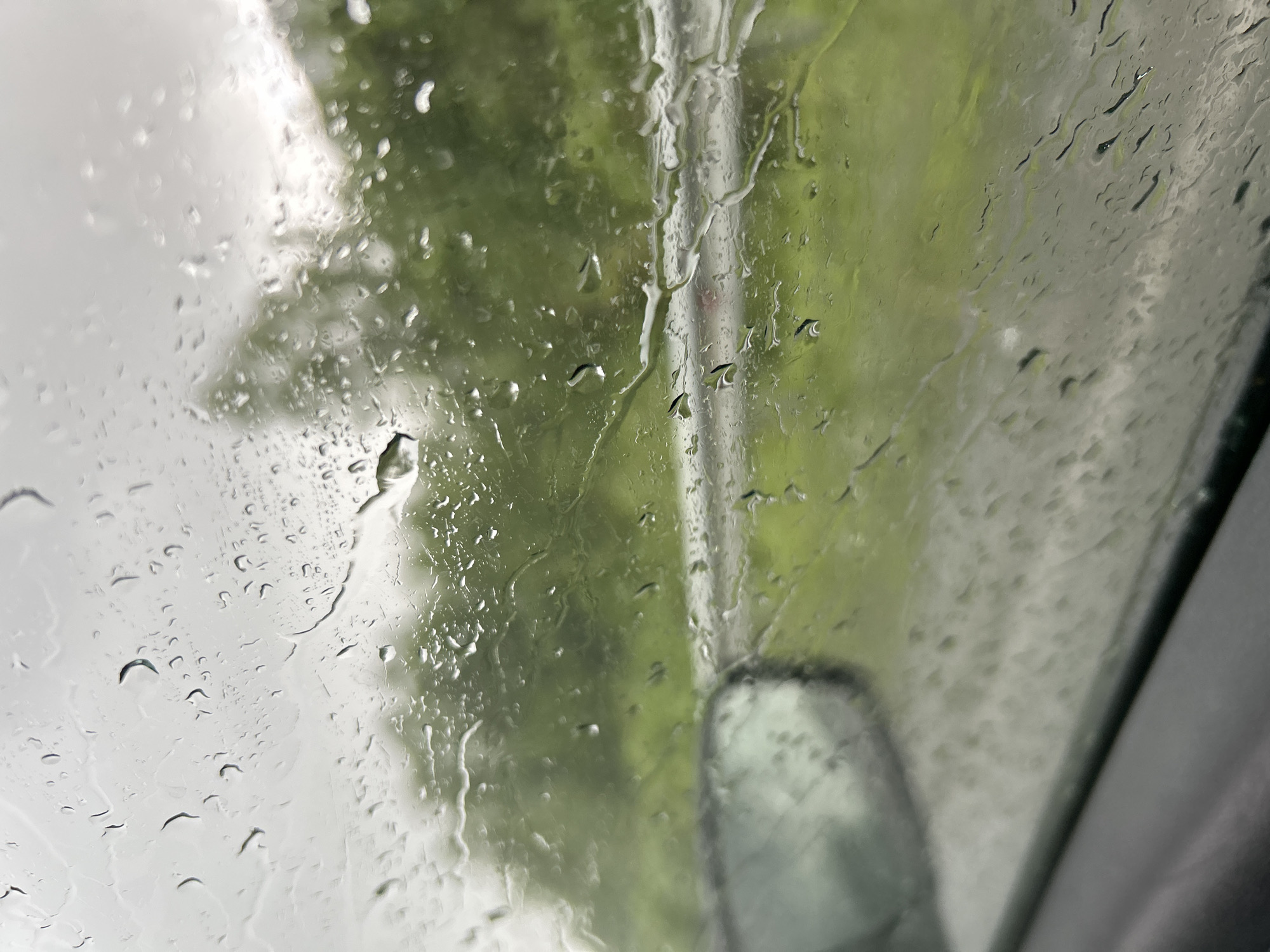 The image shows a view through a rain-streaked window, likely a car window, looking out at a blurred green and gray landscape. Water droplets of various sizes are visible on the glass, distorting the view of trees and a road. A dark, out-of-focus side mirror is visible on the left side of the frame. The sky appears overcast. The scene suggests a rainy day in an outdoor environment, possibly on a road trip or commute. The location in the city of Кулмас, Russia, is not discernible from the image itself.