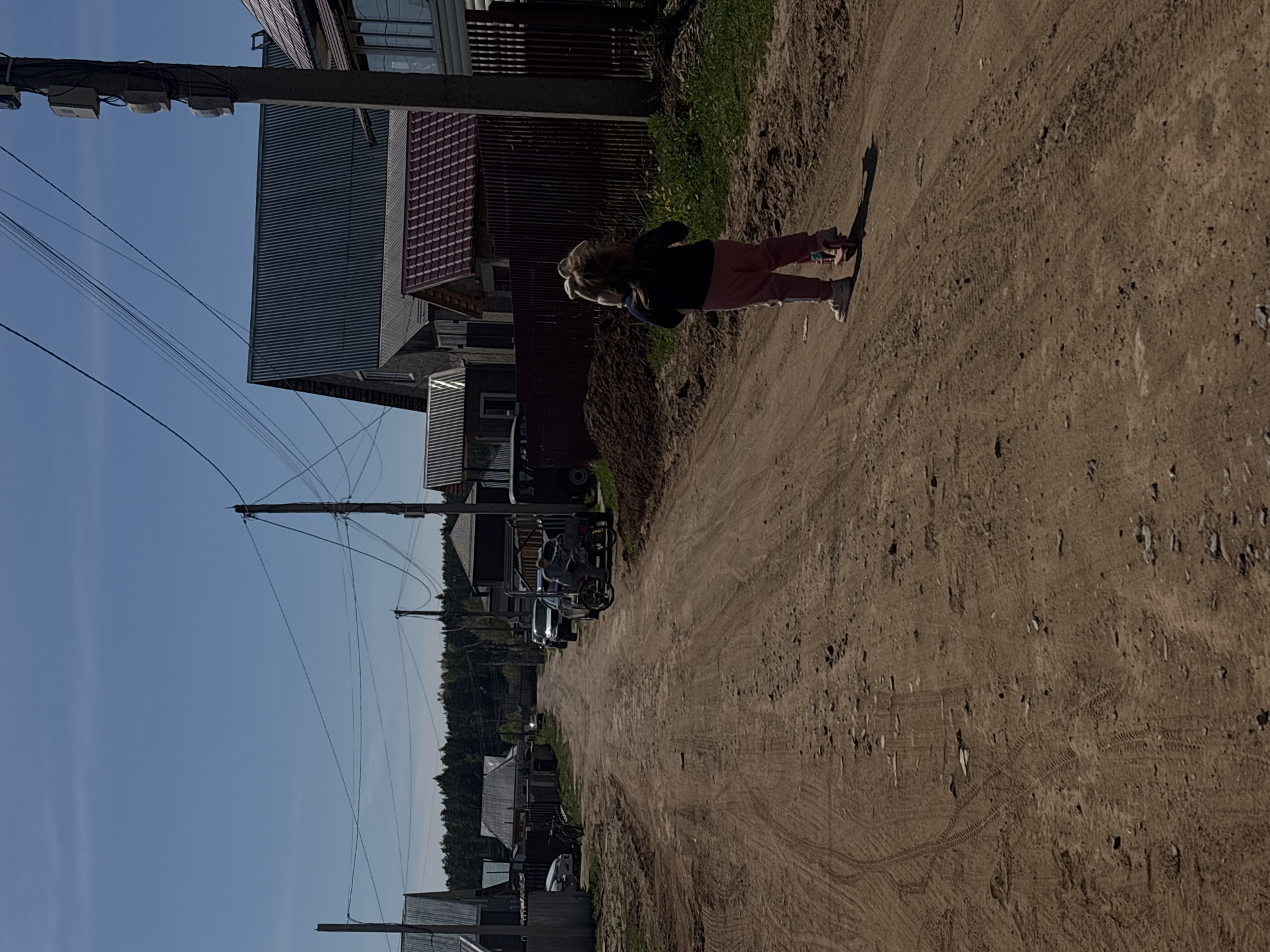A street scene in Prikamskii, Russia shows a dirt road with houses and utility poles lining either side. A person is standing on the road. Several vehicles are parked along the side of the road. The sky is clear blue.