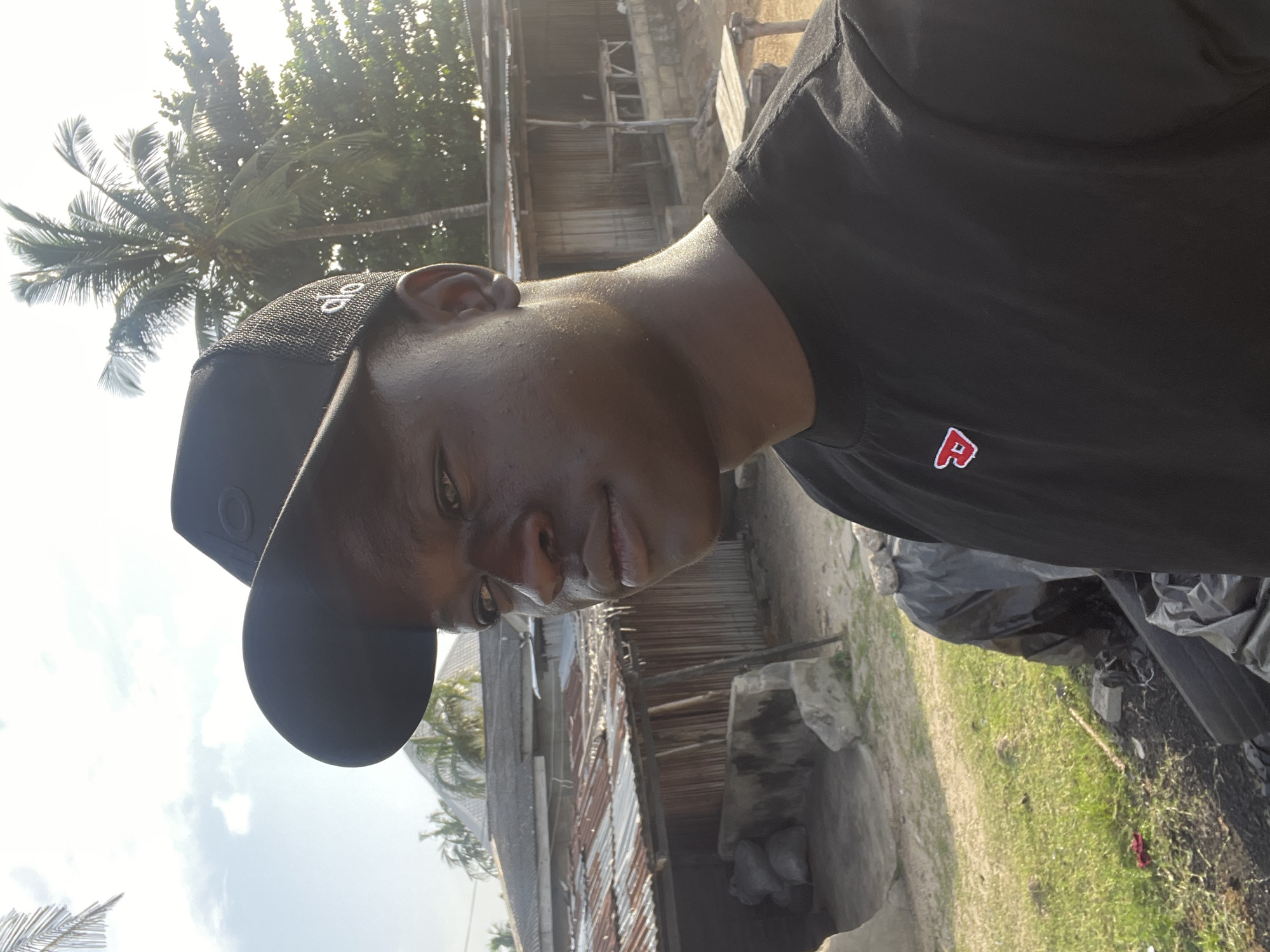 A close-up, slightly angled shot of a young Black man wearing a black t-shirt with a small red logo and a black baseball cap with white lettering. He appears to be outdoors in a sunny environment. The background includes a building with corrugated metal roofing, possibly residential, suggesting a less urban or developed area. Palm trees are visible in the background, further suggesting a tropical or coastal environment. The setting is the city of Badagry, Nigeria. There is garbage.