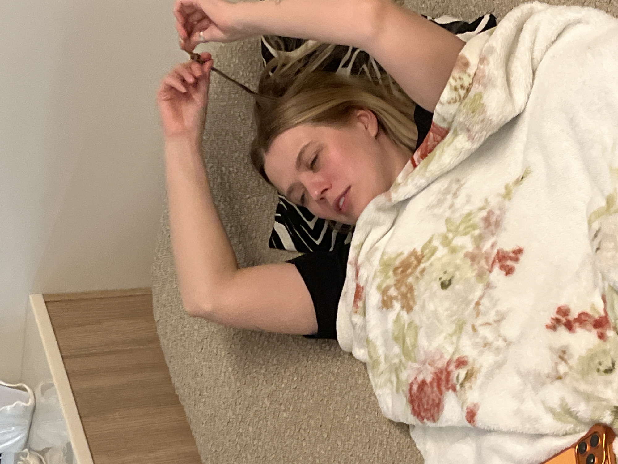 A woman is lying on a couch or bed, seemingly asleep. She is partially covered with a floral-patterned blanket. One arm is raised, and she appears to be manipulating a strand of her own hair. She has light skin and blonde hair. The location and city of Рубцово, Russia are not determinable based on the visual information in this image.