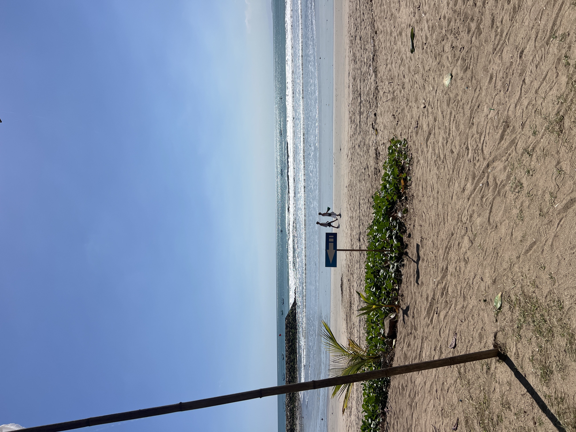 In Kuta, Indonesia, a sandy beach stretches towards the ocean under a clear blue sky. The gentle waves of the ocean create white lines of foam as they break on the shore. A breakwater, constructed with rocks, extends into the water, with several fishing boats anchored beyond it. Two individuals are walking on the wet sand near the waterline. A blue directional sign with a white arrow points to the left. In the foreground, low-lying green vegetation grows in the sand, with a tall wooden pole casting a shadow. The sand itself is textured with ripples and scattered debris.