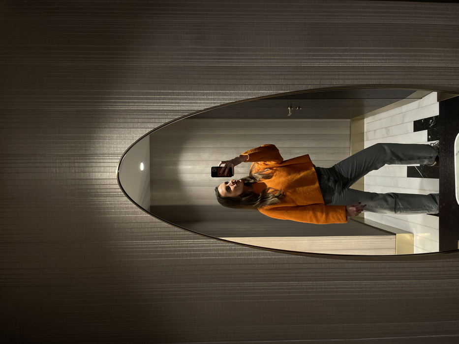A woman in an orange cropped jacket and gray jeans takes a selfie in an oval mirror. The mirror reflects a bathroom with white and black marble floors, light beige walls and recessed lighting. The surrounding wall outside the mirror has a horizontally striped texture. The reflection shows the woman holding her phone up to capture the image.