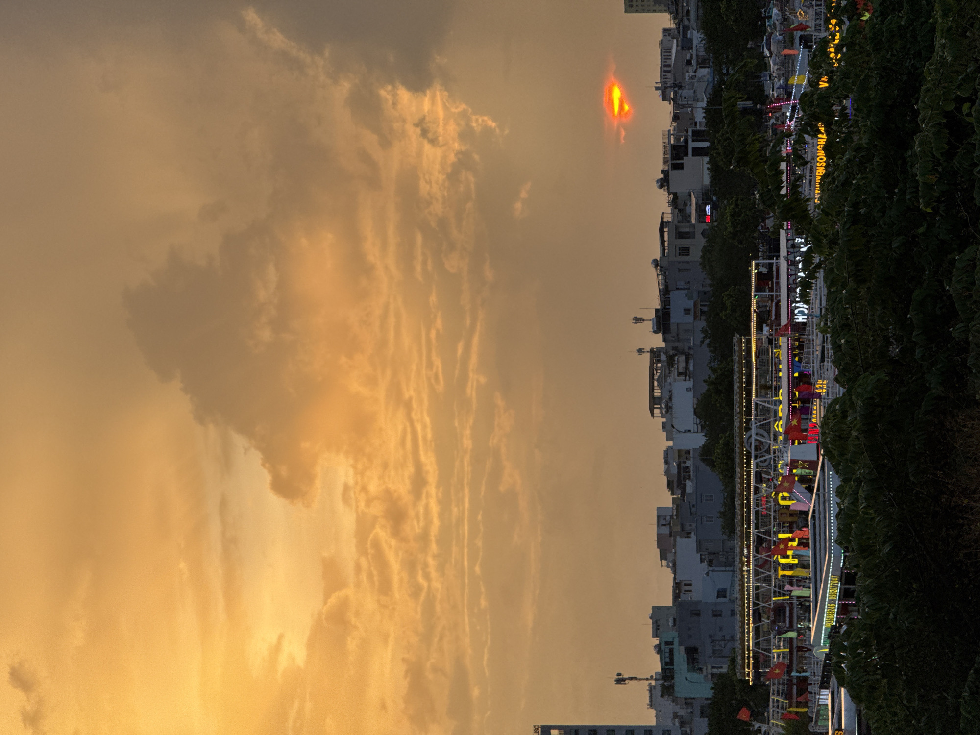 This image captures a sunset over the city of Đà Nẵng, Vietnam, likely viewed from a higher vantage point overlooking the Han River. The sky is dominated by warm, orange and yellow hues, with scattered clouds creating texture and depth. The sun, a fiery red-orange orb, is partially obscured by a cloud formation in the upper right quadrant of the sky.
Below the sky, the cityscape unfolds. A cluster of buildings, varying in height and architectural style, forms the urban backdrop. Many of these structures appear to be residential or commercial buildings, with some showing signs of illumination. Telecommunication towers are visible atop some of the taller buildings.
In the foreground, several boats are docked along the riverbank, adorned with numerous colorful flags and strings of lights. These boats are likely engaged in evening tours or dining services. The reflections of the lights from the boats shimmer on the water, although the water itself is not prominently featured in this shot. Dense green foliage, possibly trees and bushes, occupies the lower portion of the image, framing the view of the city and the river. The overall atmosphere is serene, with the vibrant colors of the sunset contrasting with the urban landscape and lush greenery.