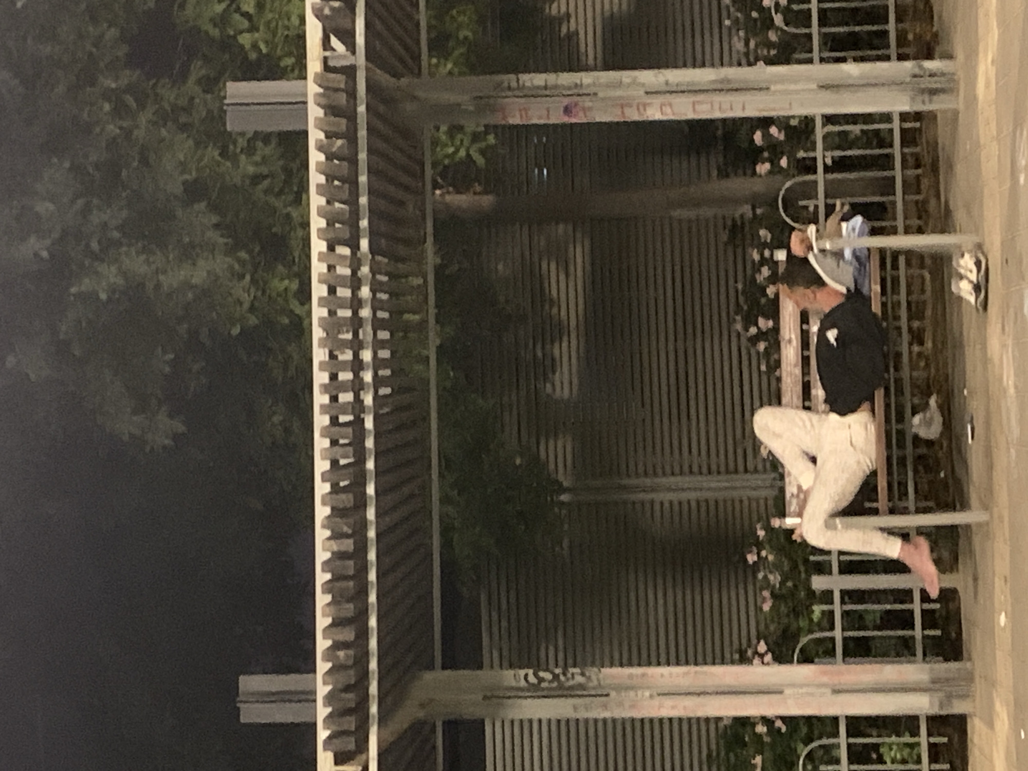 The image shows a person sitting barefoot on a bench beneath a pergola-like structure in an urban environment. The person, wearing a dark-colored long-sleeved shirt and light-colored pants, is seated with their legs extended. The structure appears to be located adjacent to a vertical slatted wall with climbing plants and flowers at its base. The left side of the image is dark, suggesting nighttime. The location is likely within the city of Tel Aviv, Israel.