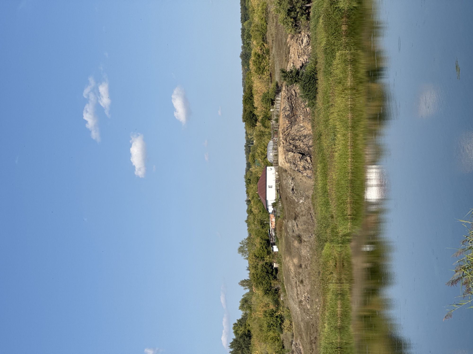 This is a vertical, eye-level, medium shot of a rural landscape with a body of water in the foreground. The sky is clear blue with a few scattered white clouds. In the middle ground, there is a gentle, grassy hill leading up to a small white house with a reddish-brown roof. The house is situated on the crest of the hill, surrounded by some trees and a fence. The hill itself is rocky in places and covered in dry grass and vegetation. The background is a dense line of trees with foliage in various shades of green and yellow, suggesting a late summer or early autumn setting. The body of water in the foreground is calm, reflecting the sky and the house on the hill. Reeds and tall grass grow along the edge of the water. The scene appears to be in a rural area, possibly the outskirts of Glubokoe, Kazakhstan, given the presence of a simple dwelling and natural landscape elements. There are no people visible in the image.