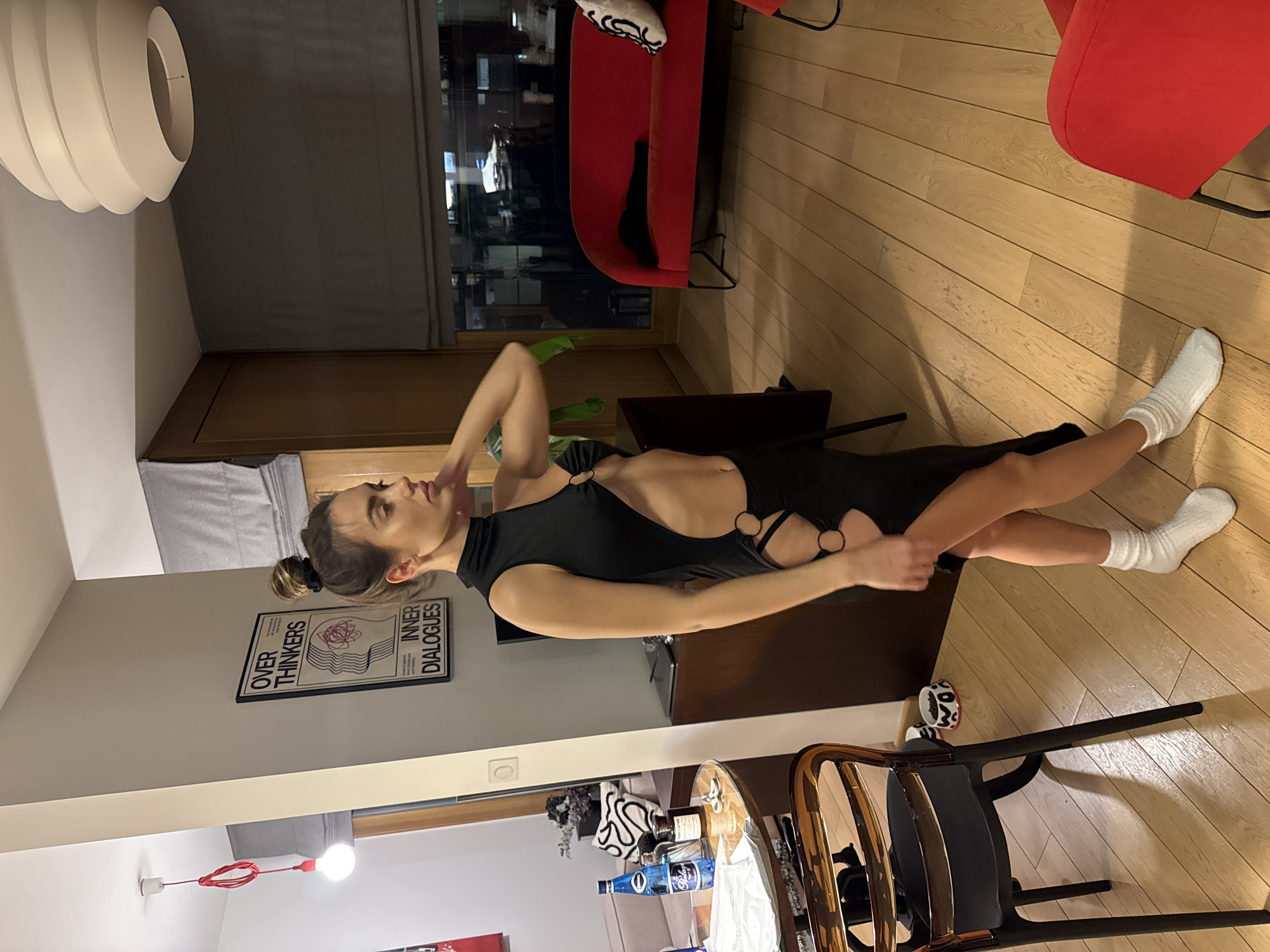 A woman stands indoors, wearing a black dress with cut-out sections and white socks. She's posing with one hand raised to her neck. The setting appears to be an apartment or living space, with hardwood floors and neutral-colored walls. There's a red sofa in the background. The woman is near a table with a gold-colored chair and various objects on it, including bottles and a white cloth.
On the wall, there is a framed poster with the words "OVER THINKERS INNER DIALOGUES." There is a pendant lamp hanging from the ceiling. The window shows a cityscape at night, which based on visual cues such as architectural styles and urban layout, is likely Warsaw, Poland.