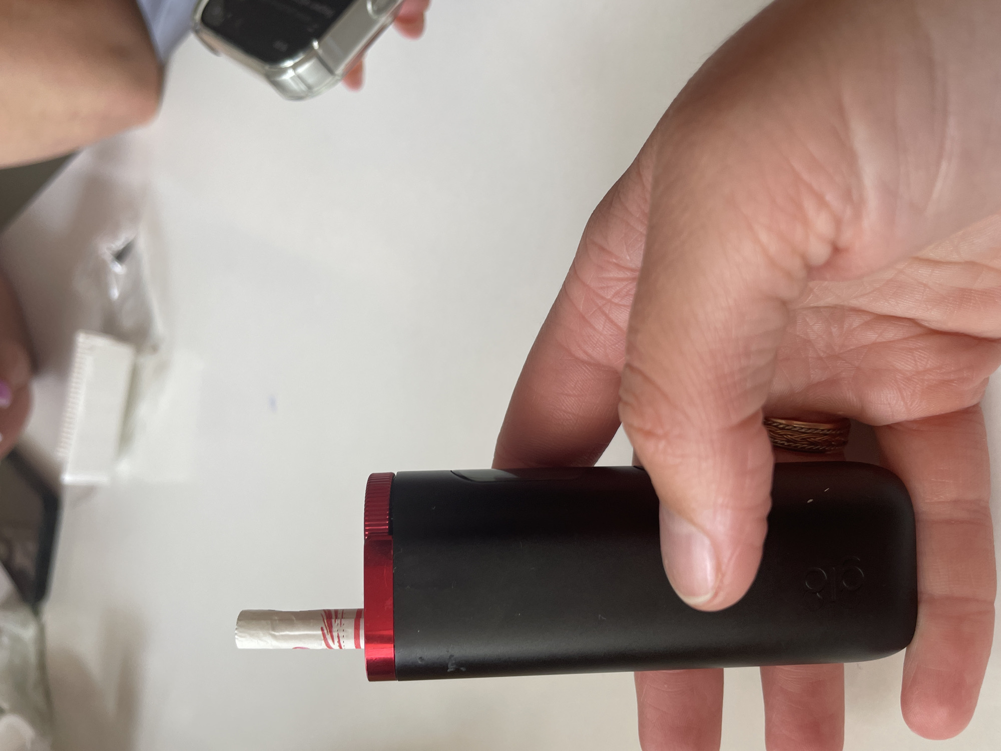 A person's hand holds a black electronic device with a red rim. A white cigarette with red stripes is inserted into the top of the device. The device has a logo on the bottom that appears to be "816". The background is a white tabletop with some blurry objects, including a phone and what appears to be packaging. Another hand is visible on the right side of the image, holding a silver-colored mobile phone.
