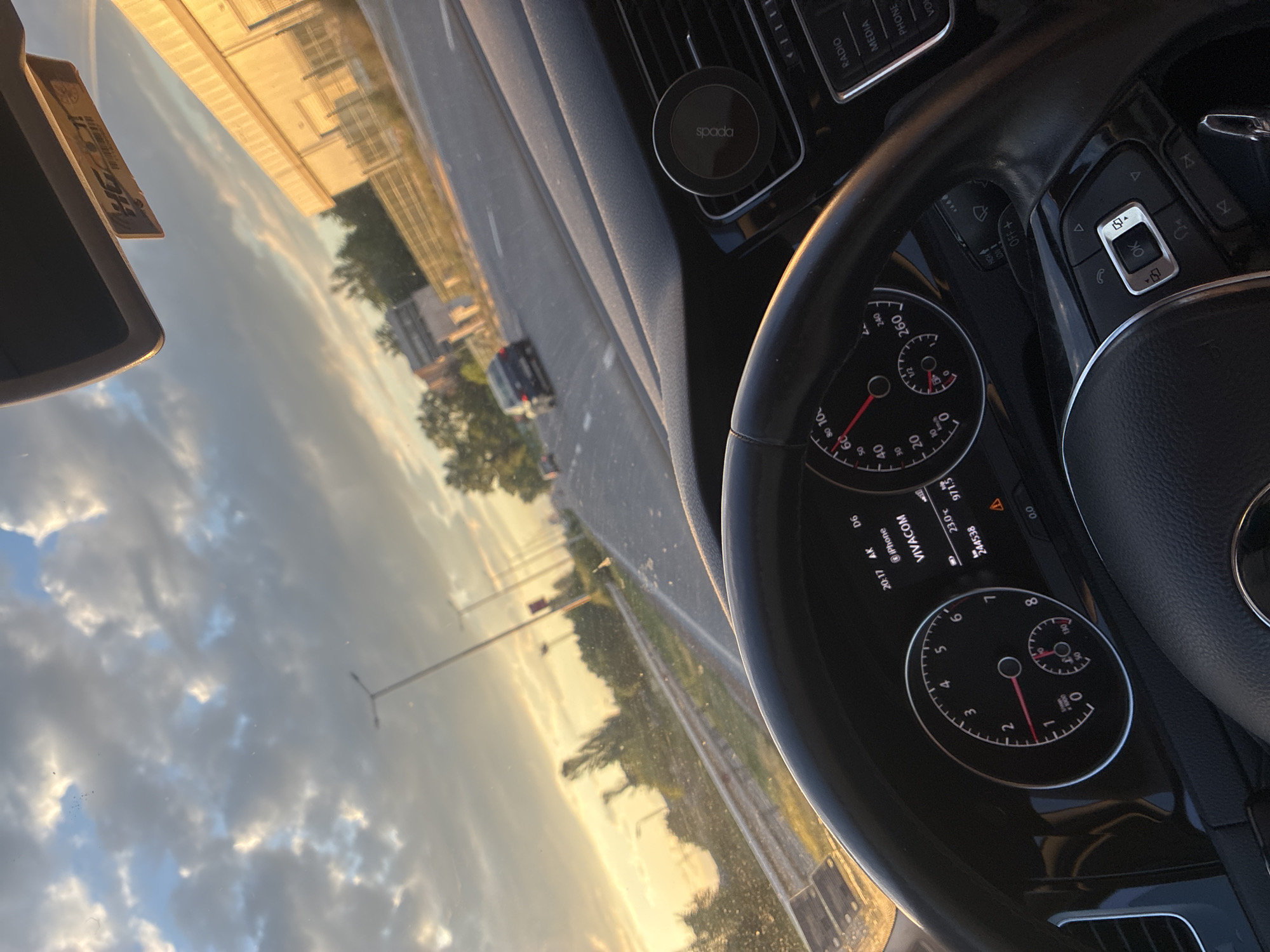 The image shows the interior of a car, specifically the dashboard, steering wheel, and windshield view. The car appears to be in motion, as indicated by the blurred scenery outside. The dashboard includes the speedometer, fuel gauge, and other information displays. A phone mount labeled "spada" is attached to the air vent. The windshield provides a view of a road with buildings, trees, and streetlights under a cloudy sky. The time displayed on the dashboard is "20:17 AK."