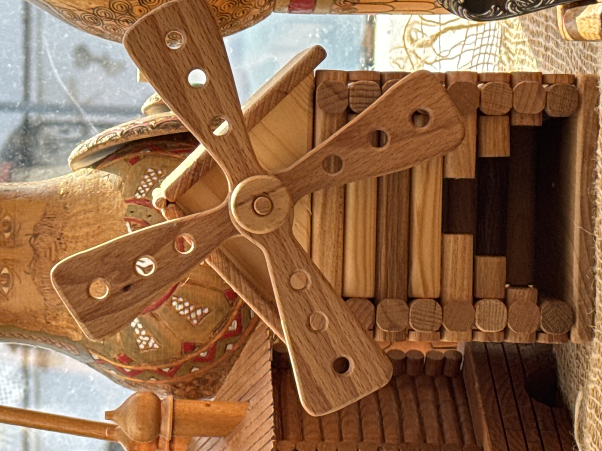 The image depicts a close-up of a wooden toy windmill placed beside an ornately decorated wooden container. The windmill features a four-bladed rotor with evenly spaced holes on each blade, and it is mounted on a structure made of rectangular and cylindrical wooden pieces. To the left of the windmill, a wooden container displays intricate painted designs in earthy tones. The objects appear to be positioned near a window, with diffused light filtering through, and the background hints at the presence of other decorative items. The location is identified as Unknown, Unknown.