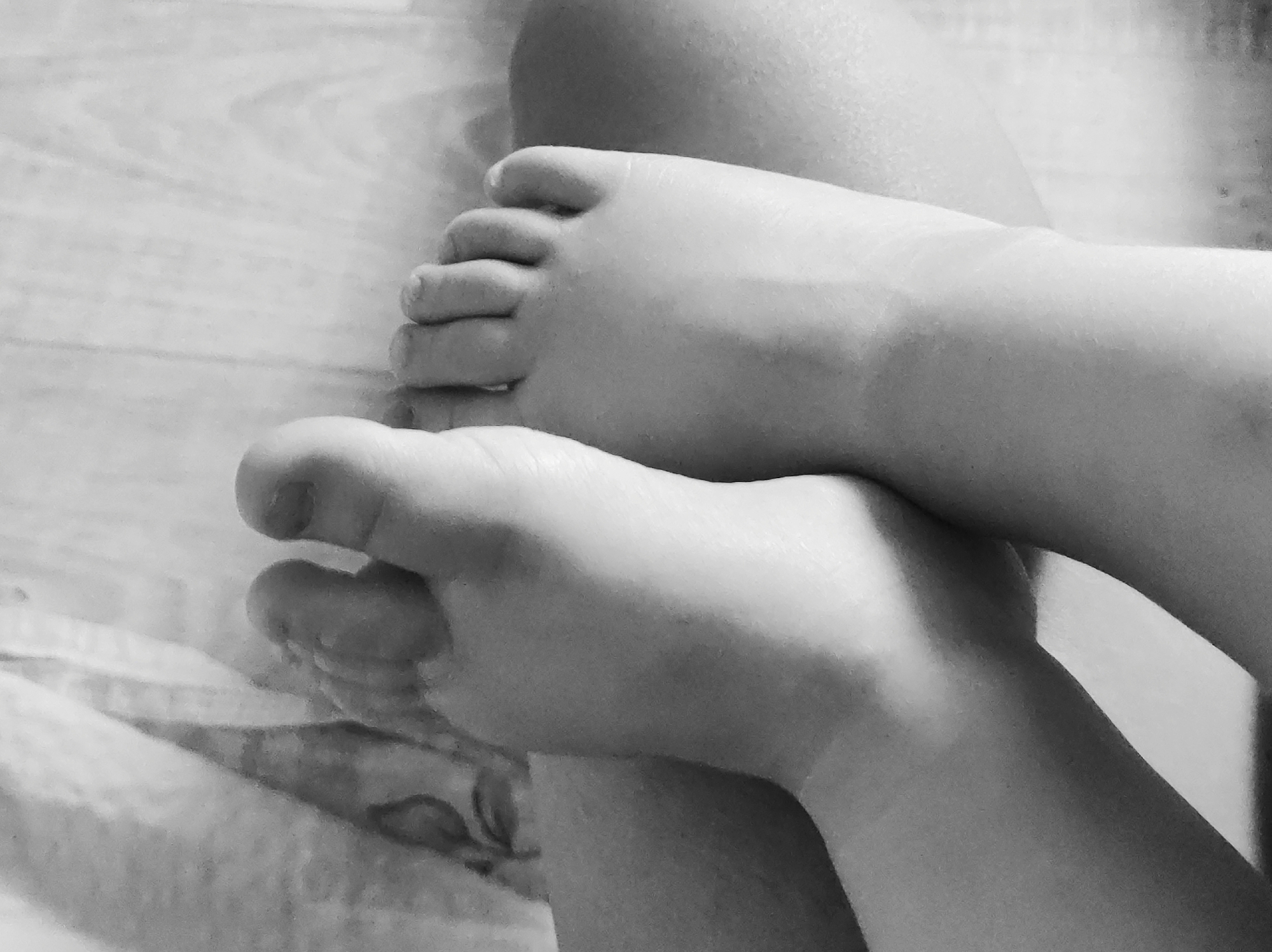 A black and white, close-up photograph features two bare feet. The feet are positioned one over the other, with toes visible on both. The skin appears smooth and soft. The background consists of a wooden surface, possibly a floor. There is also a blurry pattern on the bottom left corner, might be bedsheet. The location is unidentified.