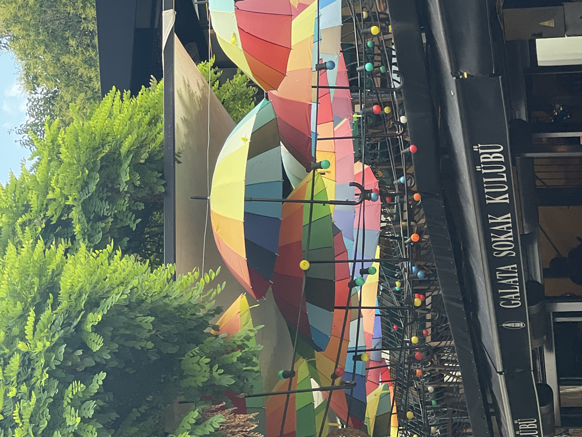 The image depicts a street scene in the Beyoğlu district of Istanbul, Turkey. Above a black awning with white text that reads "GALATA SOKAK KULÜBÜ", a collection of colorful umbrellas are suspended, creating a vibrant canopy. These umbrellas are arranged in layers and are a mix of solid colors and multi-colored segments. Interspersed with the umbrellas are string lights, featuring individual bulbs in various colors such as red, yellow, blue, green, and white. Lush green foliage from trees overhangs the scene from the upper left. Below the umbrellas and lights, the awning of a establishment is visible, with part of a name appearing as "LÜBÜ" on the left and "GALATA SOKAK KULÜBÜ" on the right. The overall atmosphere suggests an outdoor seating area for a cafe or restaurant, decorated for a festive or cheerful ambiance. The lighting indicates it is daytime, with natural light filtering through the greenery and umbrellas. There are no discernible people in the frame, but a shadowy figure is faintly visible through the glass of the establishment.