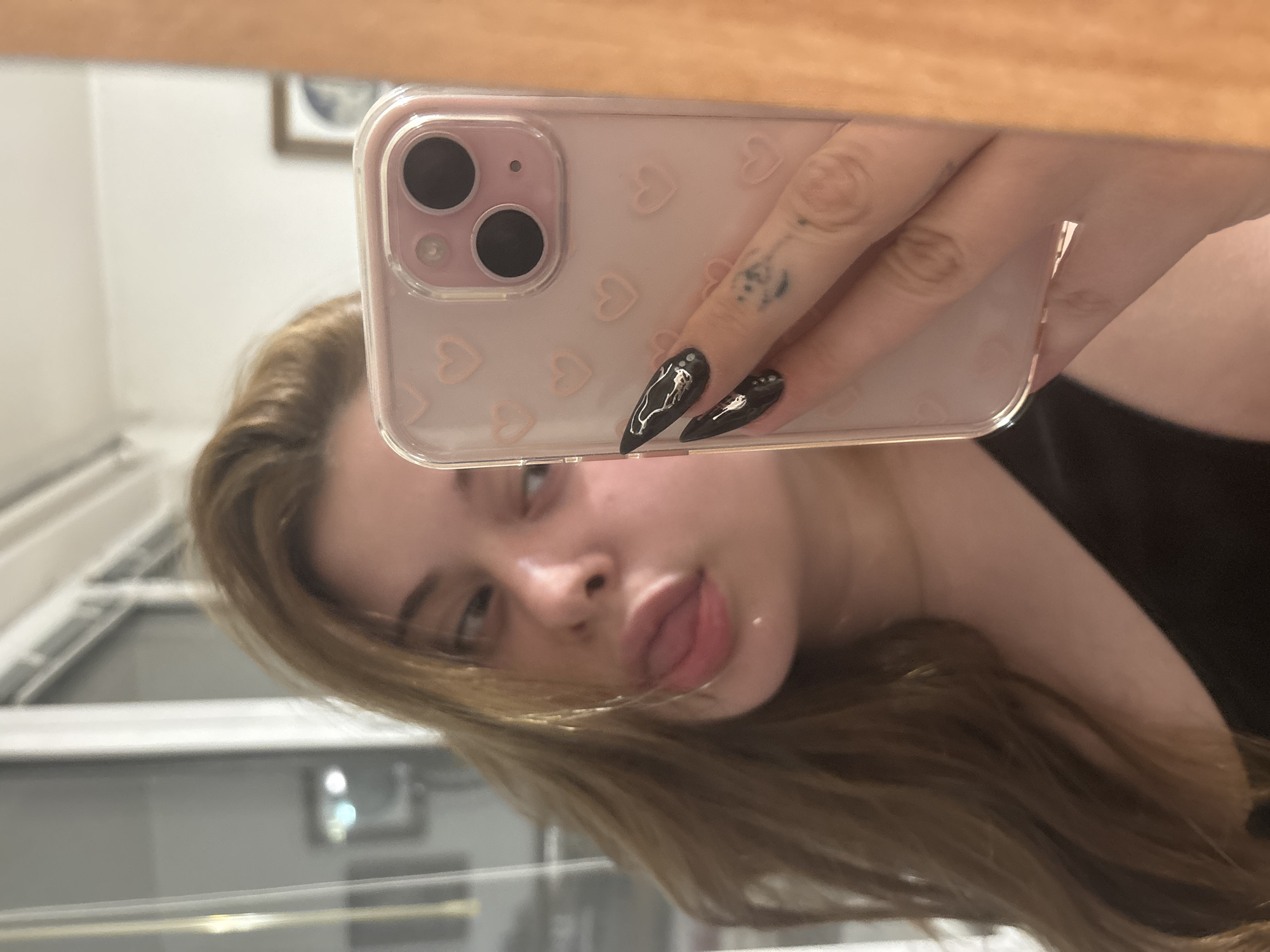 A young woman is taking a selfie in a bathroom. She is holding a pink iPhone in a case decorated with hearts. Her nails are long and black with white designs. She has a tattoo on one of her fingers. The background includes a window with blinds and a framed picture on the wall. The location is indeterminate, and cannot be confirmed to be in Troyan, Bulgaria.