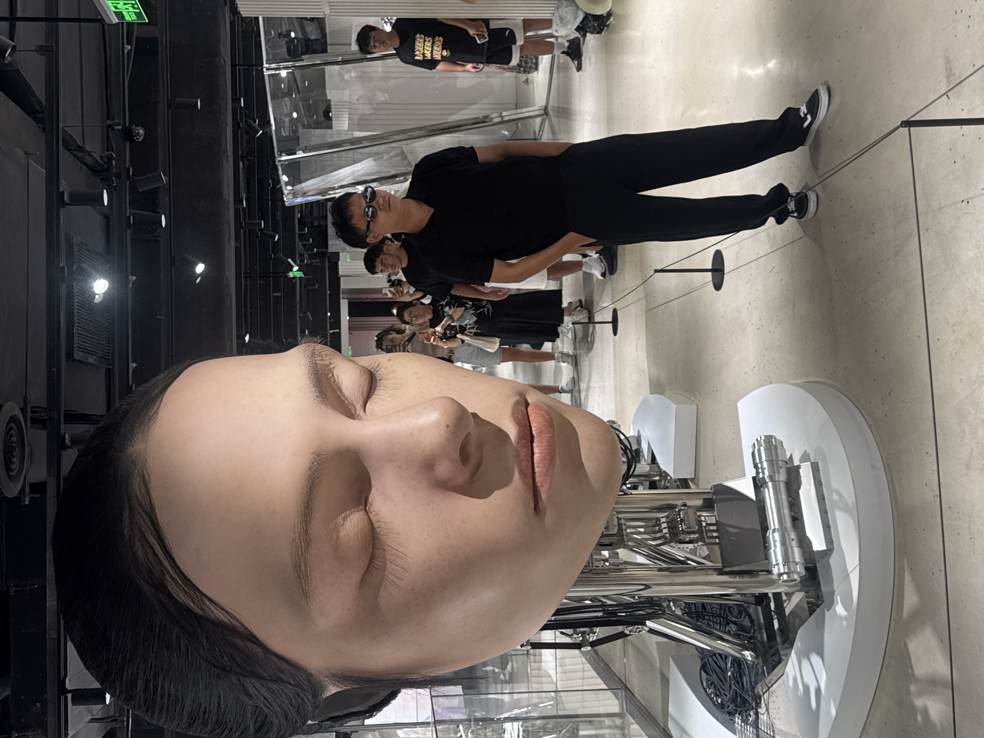 The image depicts a large, realistic sculpture of a human head, likely female, with closed eyes and rendered in a pale, lifelike skin tone, positioned on a metallic armature. The sculpture is located in a well-lit indoor space that appears to be a museum or exhibition hall. Several people are present, observing the artwork. In the foreground, a man wearing sunglasses, a black polo shirt, and black pants stands with his hands in his pockets, facing the sculpture. Behind him, other individuals, including a man holding a phone and a woman taking a photo, are also visible. The background features a modern interior with exposed ceiling structures, track lighting, and reflective surfaces, suggesting a contemporary exhibition setting. The overall environment is clean and organized, with stanchions and ropes delineating viewing areas. The lighting is bright, casting shadows that emphasize the contours of the sculpture and the people. Given the modern aesthetic and exhibition style, this scene is likely located in a contemporary art museum or a design exhibition in Shanghai, China, potentially within a district known for its cultural institutions such as the West Bund or Pudong.