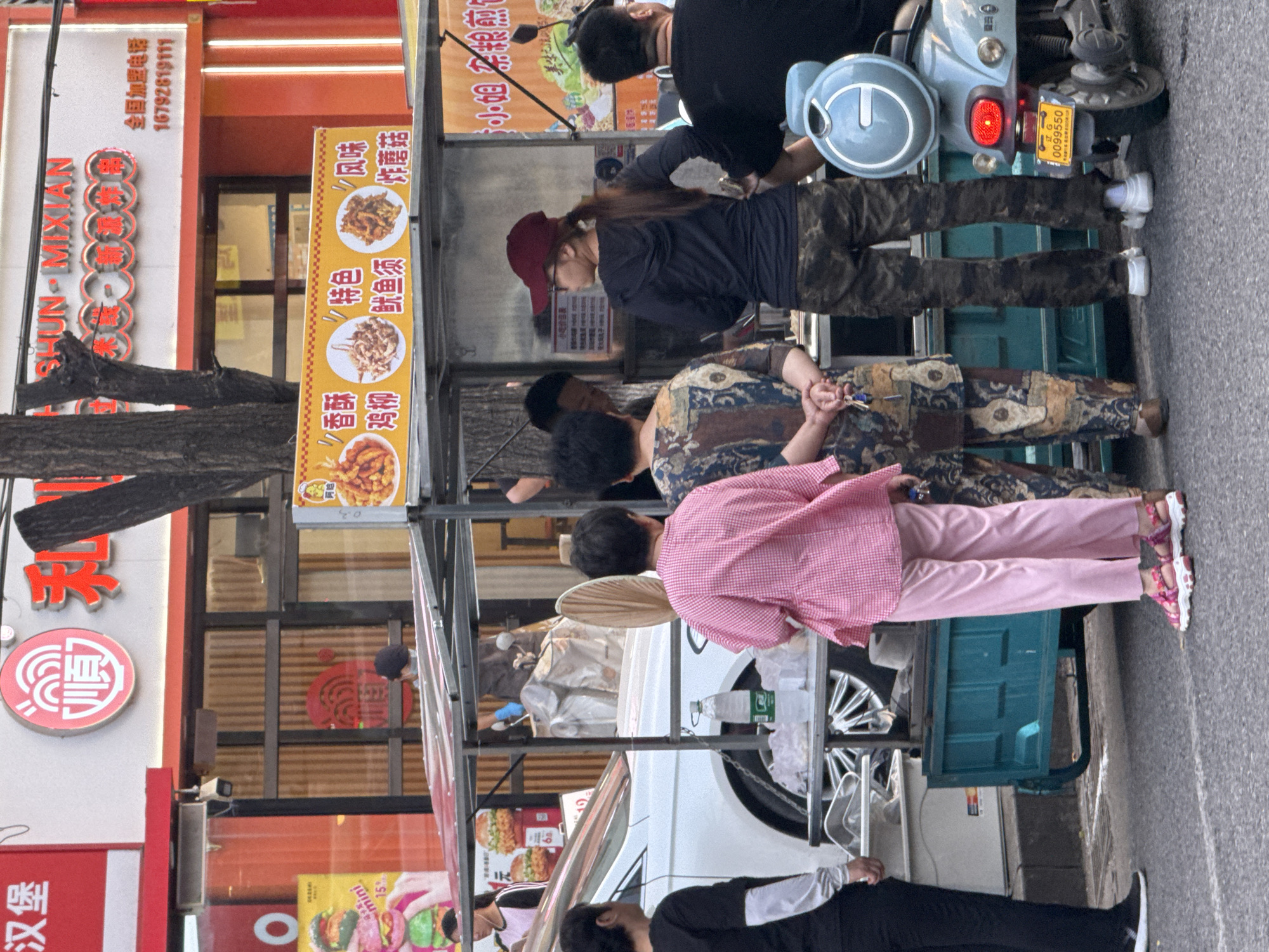 The image depicts a street scene in Zhongtun, China, with a food stall as the central focus. The food stall appears to be mobile, with a turquoise cart base and a transparent canopy. Several people are present, some interacting with the stall and others standing nearby.
In the foreground, a person wearing a pink checkered shirt and pink pants stands with their back to the camera, facing the food stall. To their left, another person in a black long-sleeved shirt and black pants stands with their left side visible. Behind the food stall, several individuals are partially obscured. One person, wearing a dark hoodie and camouflage pants, stands looking towards the stall. To their left, a person in a patterned dress is visible.
The food stall itself displays a variety of fried foods, indicated by images and text on yellow banners. Some of the visible text suggests items like "香酥鸡柳" (crispy chicken strips), "特色鱿鱼须" (specialty squid tentacles), and "风味炸蘑菇" (flavored fried mushrooms). A white car is parked to the left of the food stall. In the background, there is a building with signage, including what appears to be a restaurant. The street is paved, and street lights are visible, suggesting an urban environment. A blue scooter with a license plate reading "正 G 0099550" is parked to the right of the food stall, with its brake light illuminated. The overall scene suggests a bustling street with food vendors and pedestrian activity.
