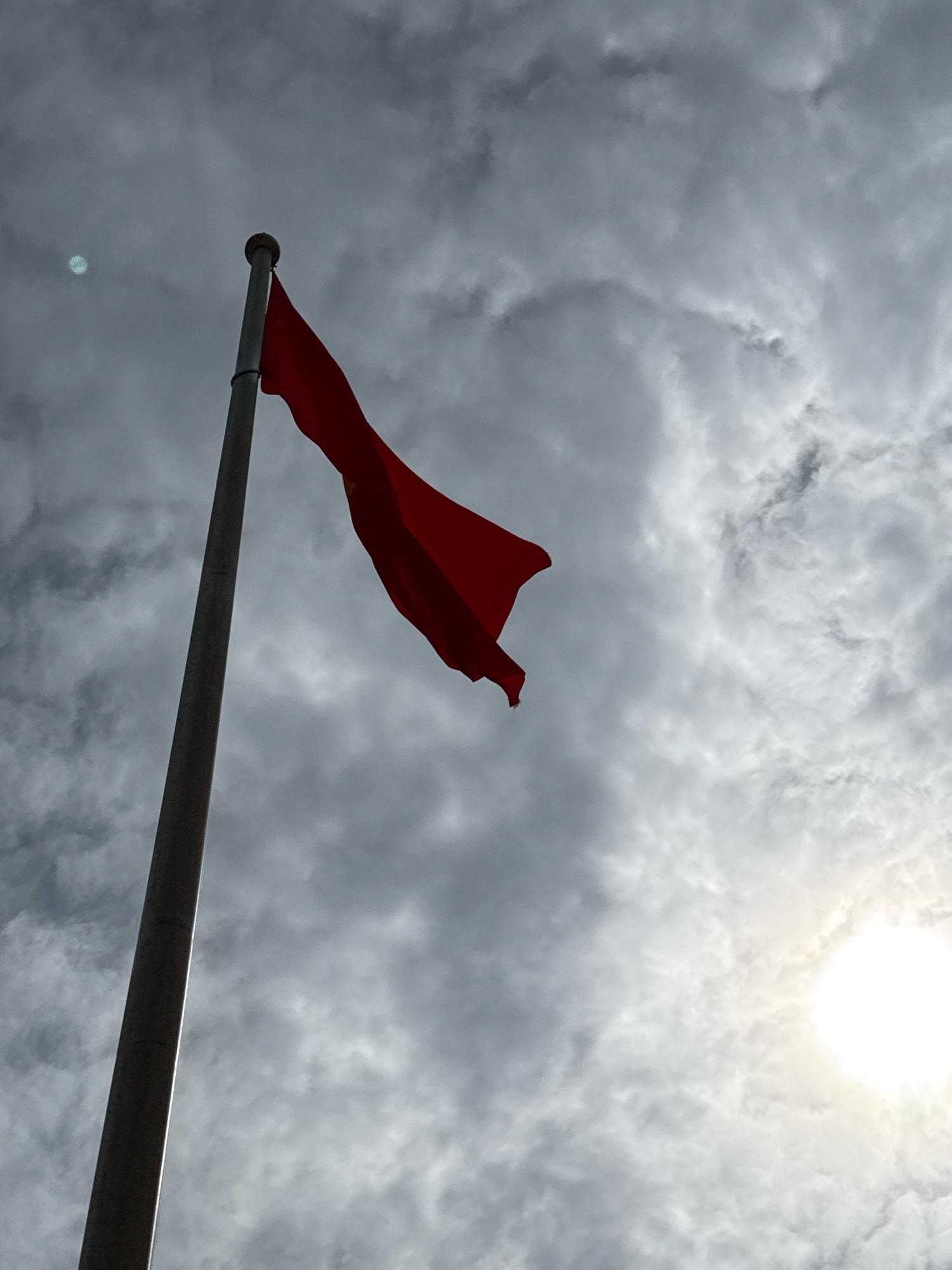 A low-angle, close-up shot captures a solitary, red flag billowing majestically against a dramatic, cloudy sky. The flag, affixed to a tall, slender flagpole, is angled towards the viewer's right, its fabric caught in an unseen wind, creating dynamic curves and folds. The flagpole, a metallic gray, stands vertically on the left side of the frame, its tip featuring a rounded, light-colored cap. The sky above is a tapestry of gray clouds, some dense and dark, others lighter and wispy, hinting at an overcast or approaching storm. The sun is visible in the upper right quadrant, a bright, hazy orb peeking through the clouds, casting a soft, diffused light. There are no people or other objects visible in the frame. The image appears to be taken from an outdoor location, possibly a park, government building, or a public square in Nha Trang, Vietnam, given the context.