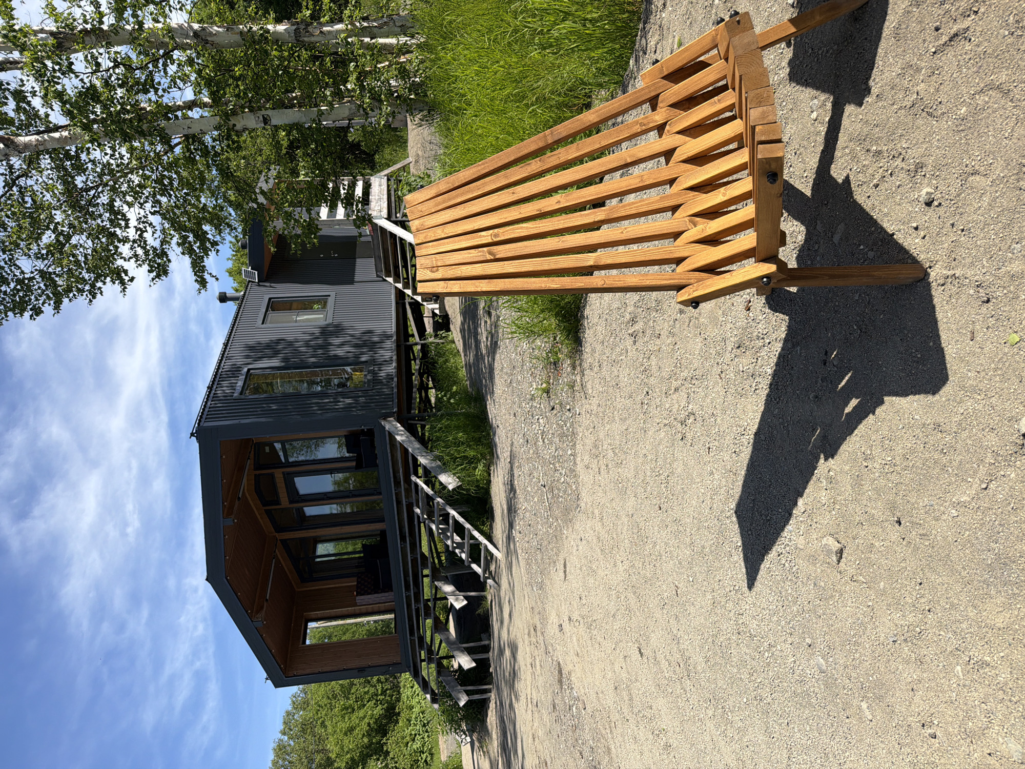 The image depicts an outdoor scene in a rural or semi-rural area, possibly near a body of water, given the presence of grass and trees. In the foreground, a wooden lounge chair is positioned on a gravel surface. The chair is made of light-colored wood, with a slatted design that allows for reclining. Its shadow is cast on the ground, indicating a sunny day.
In the midground, a modern, dark-colored house or cabin stands on stilts. The structure features large windows and a corrugated metal exterior. A wooden deck and a set of stairs lead up to the entrance. The house appears to be situated amongst trees and vegetation.
The background shows a clear blue sky with scattered white clouds and more trees. The overall impression is one of a serene, possibly vacation-like setting. The location is stated to be in the city of Novy, Russia.